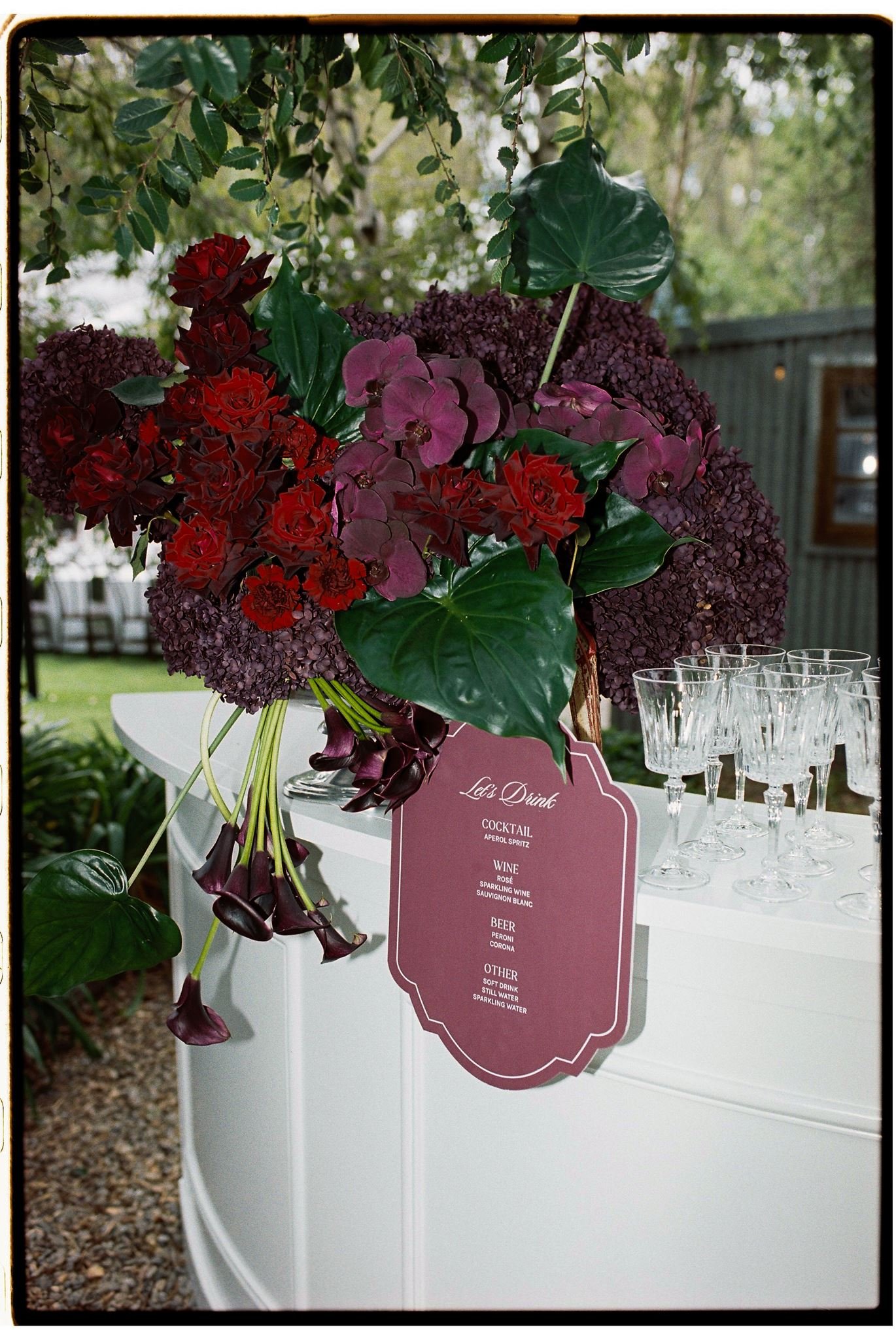 A floral centerpiece with large green leaves, red and purple flowers, and pink hydrangeas on a white table at an outdoor event. A pink menu card is visible, listing drinks and other beverages.