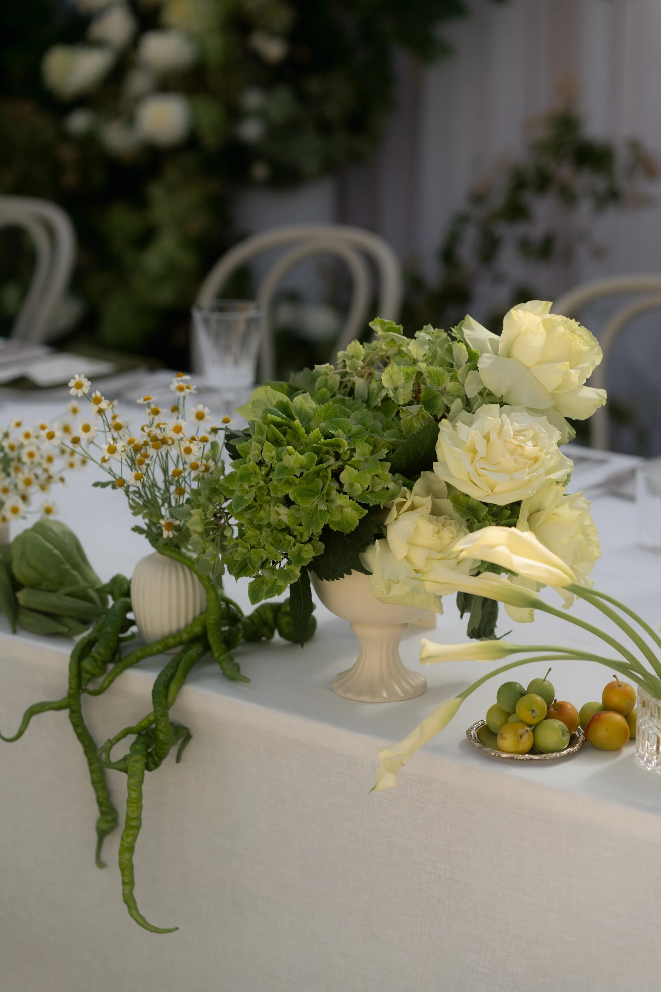 A floral table centerpiece with white roses, green foliage, and small white daisies in a ceramic vase on a white tablecloth, with a backdrop of green plants.
