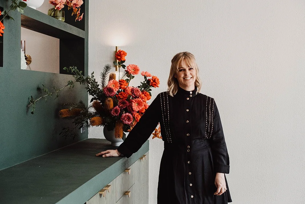 A smiling woman with blonde hair in a black dress standing next to a large bouquet of colorful flowers on a green countertop inside a room with white walls.