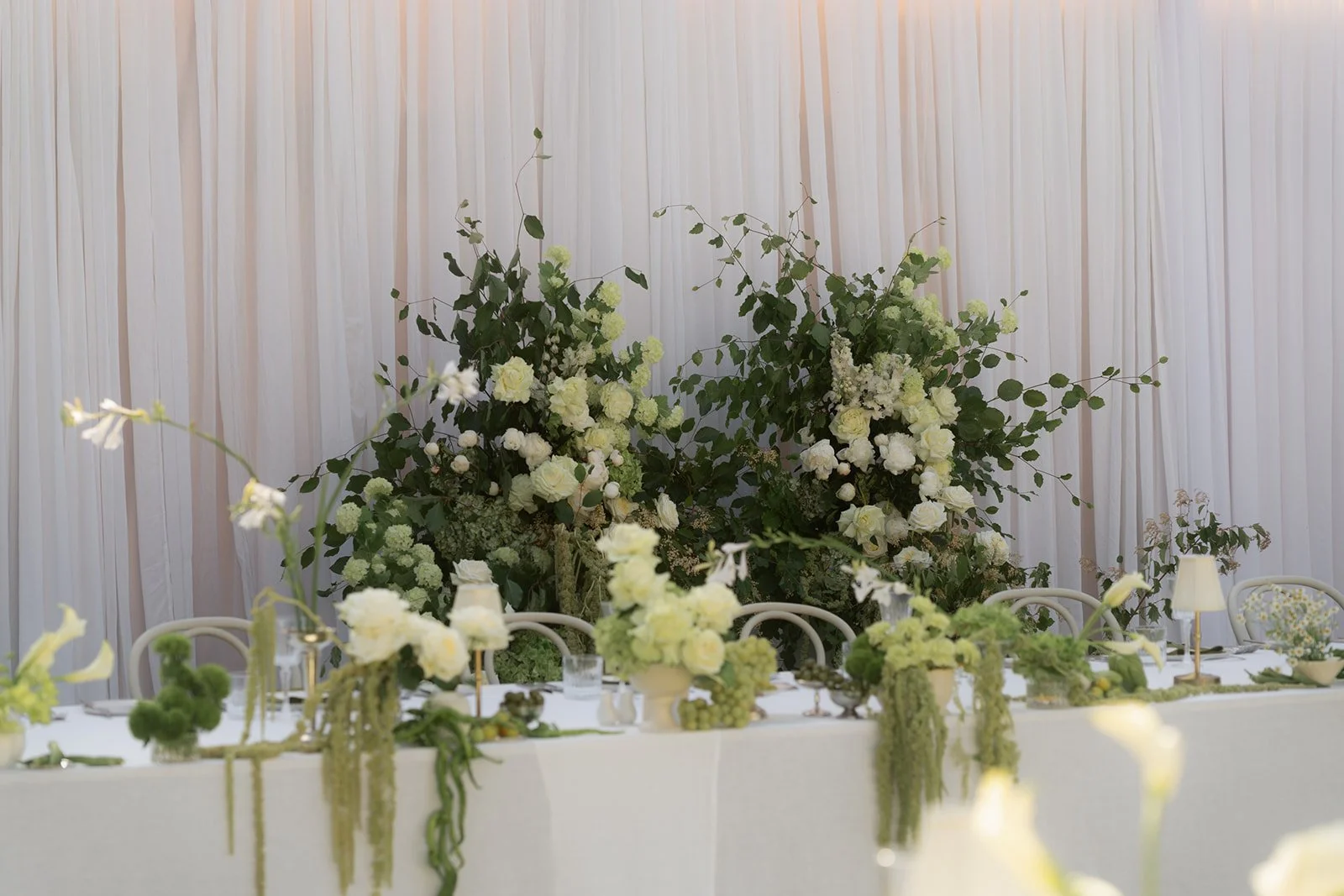A table decorated with white flowers and greenery, with a large floral arrangement in the background against white curtains, set for a wedding or special occasion.