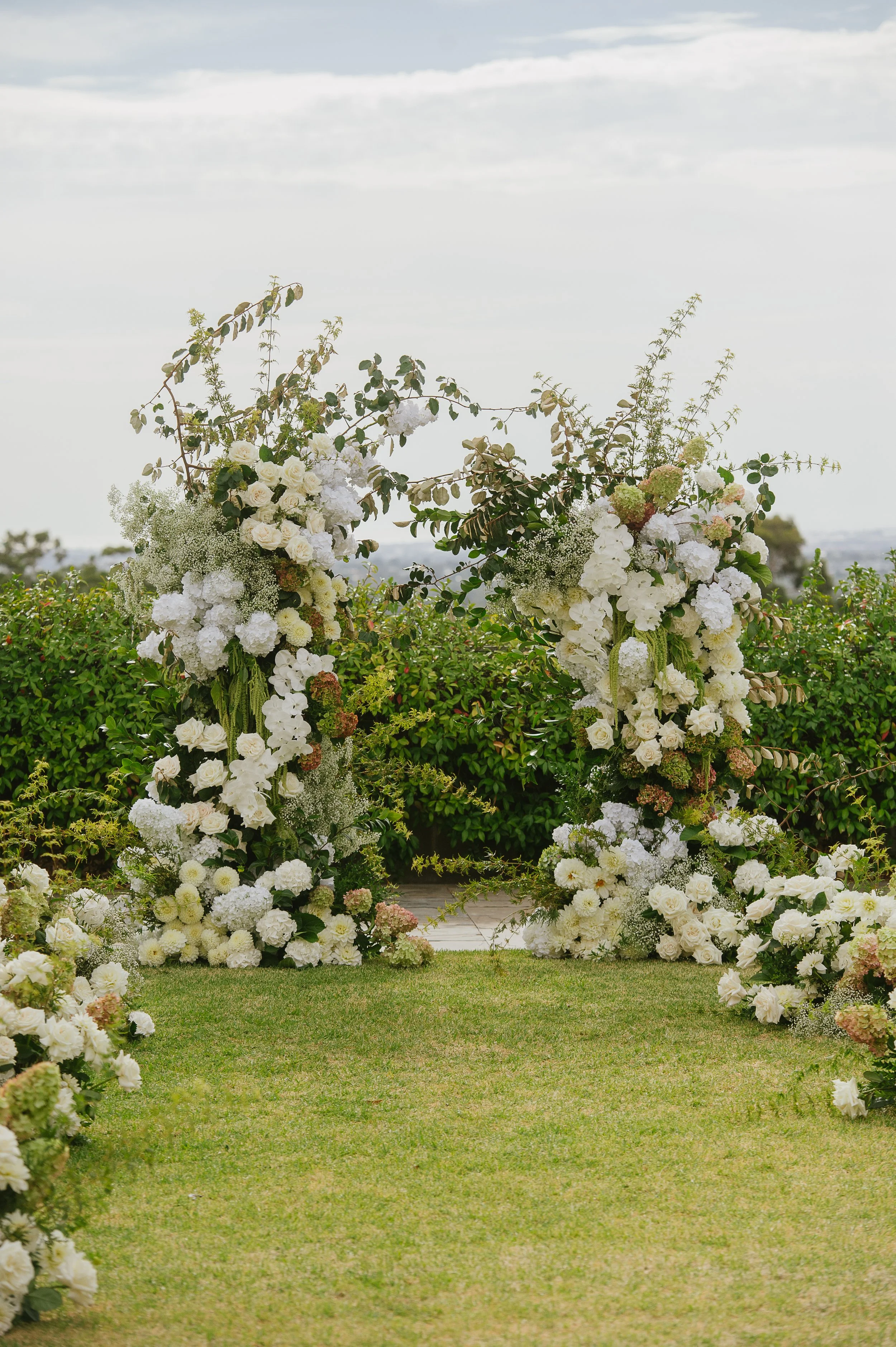 Floral wedding arch made of white roses, hydrangeas, and greenery on a grassy outdoor area with a cloudy sky background.