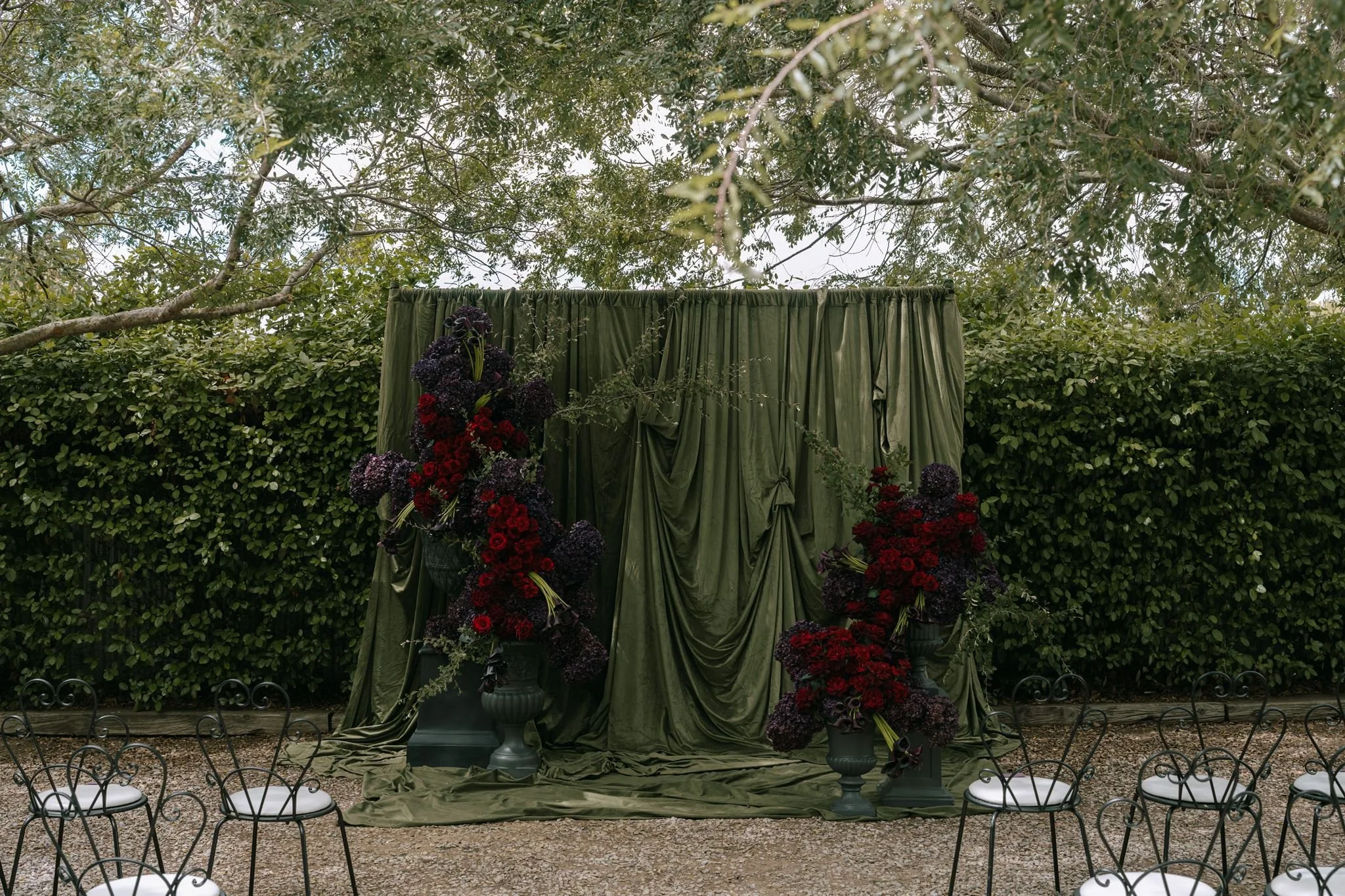 A floral arrangement with dark purple and red flowers in front of a green curtain backdrop, surrounded by lush green bushes and trees.