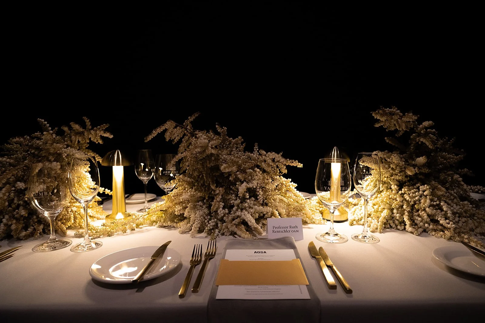 Elegant dinner table setup with white floral centerpieces, wine glasses, gold utensils, and a small table lamp, set against a dark background.