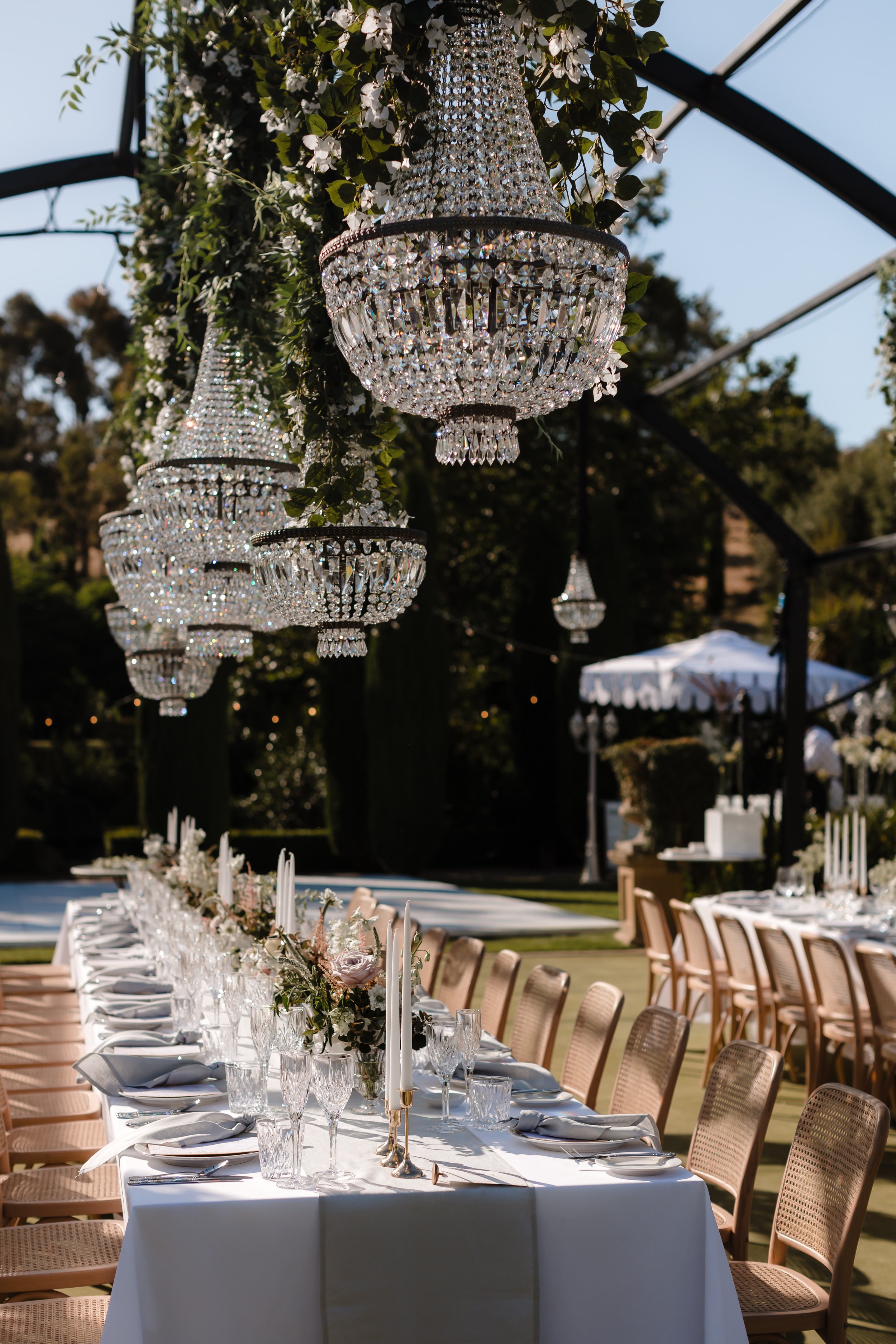 Elegant outdoor wedding reception setup with a long dining table, floral centerpieces, tall white candles, crystal chandeliers hanging from above, and a canopy pergola, under a clear sky.