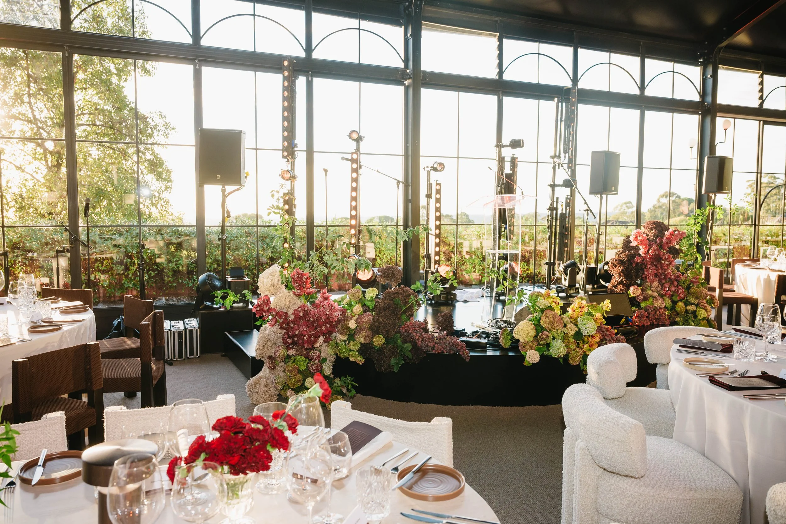 Luxury banquet hall decorated with vibrant flower arrangements in pink, white, and purple, set for an event with white and brown chairs, tableware, and a stage area with lighting and sound equipment, large windows letting in natural light.