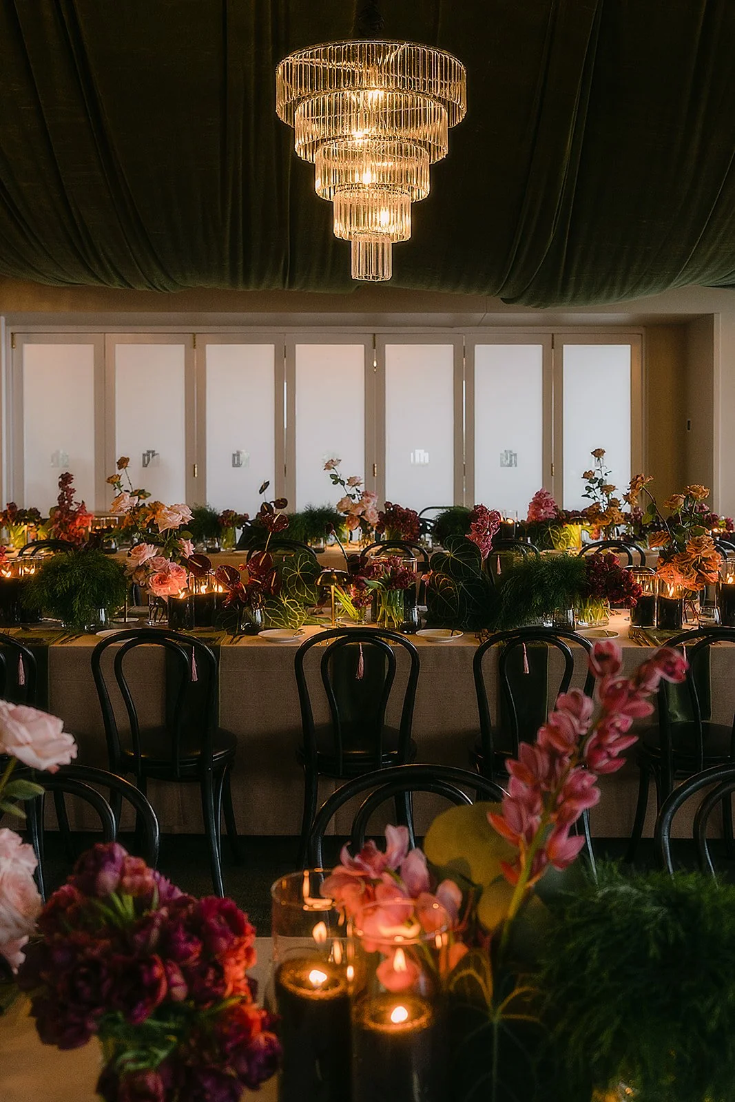 A dining room with a long table decorated with pink and burgundy floral centerpieces, small lit candles, and black chairs. There is a large chandelier hanging from a black draped ceiling above. The background has a row of white framed windows.