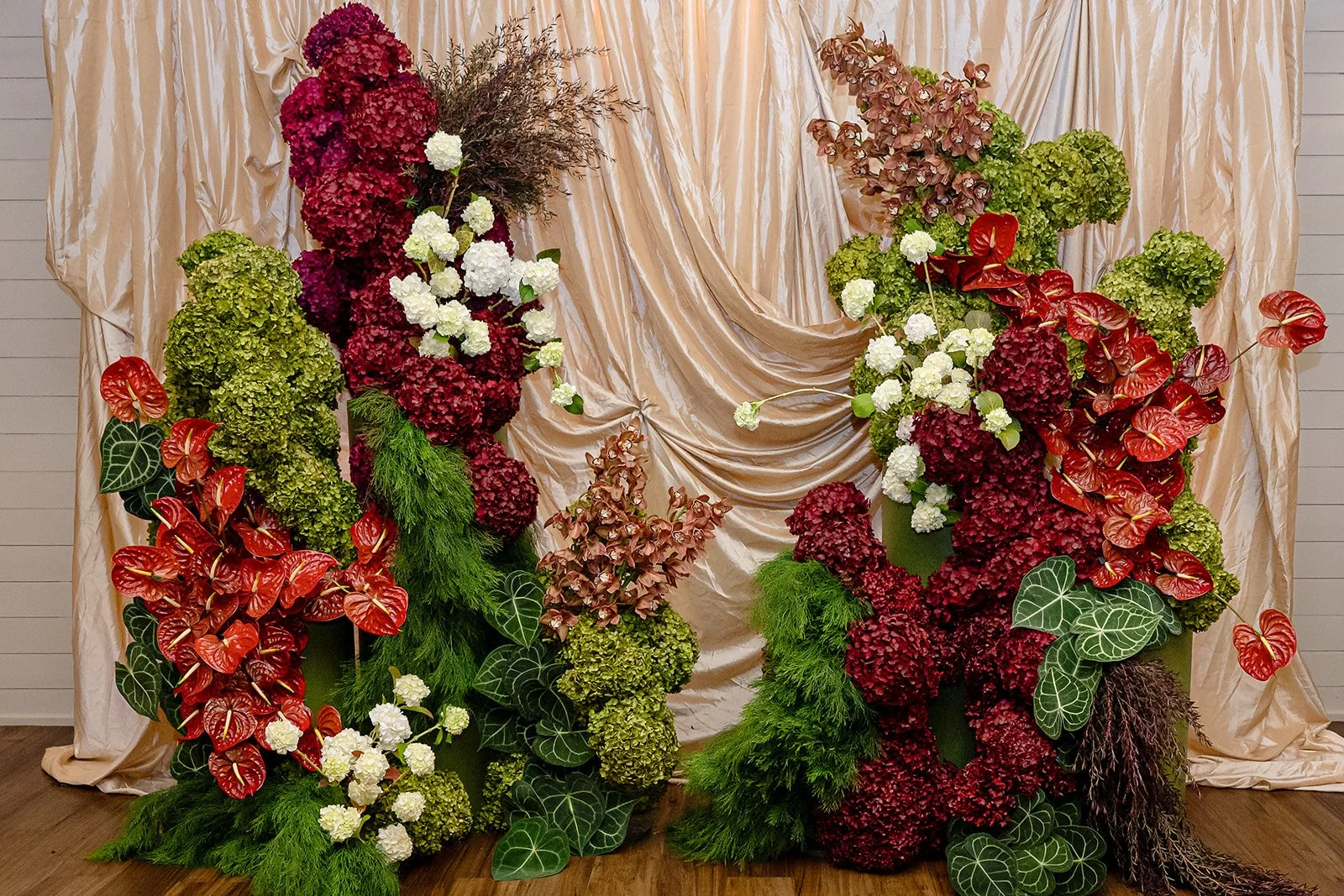A floral arrangement with various green, red, purple, and white flowers and leaves in front of a beige satin curtain backdrop.