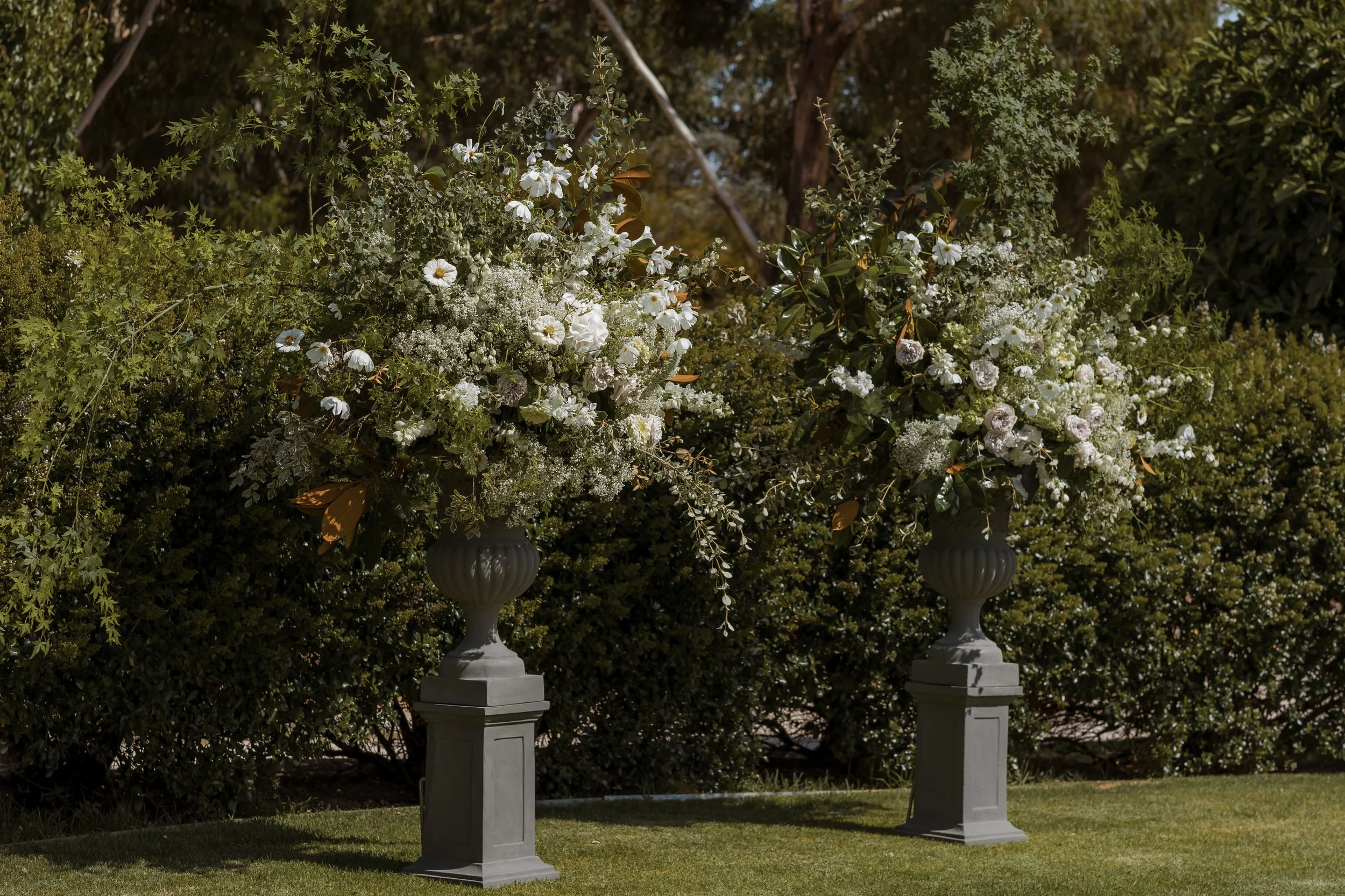 Two large floral arrangements in gray vases on gray pedestals outdoors, surrounded by green bushes and grass.