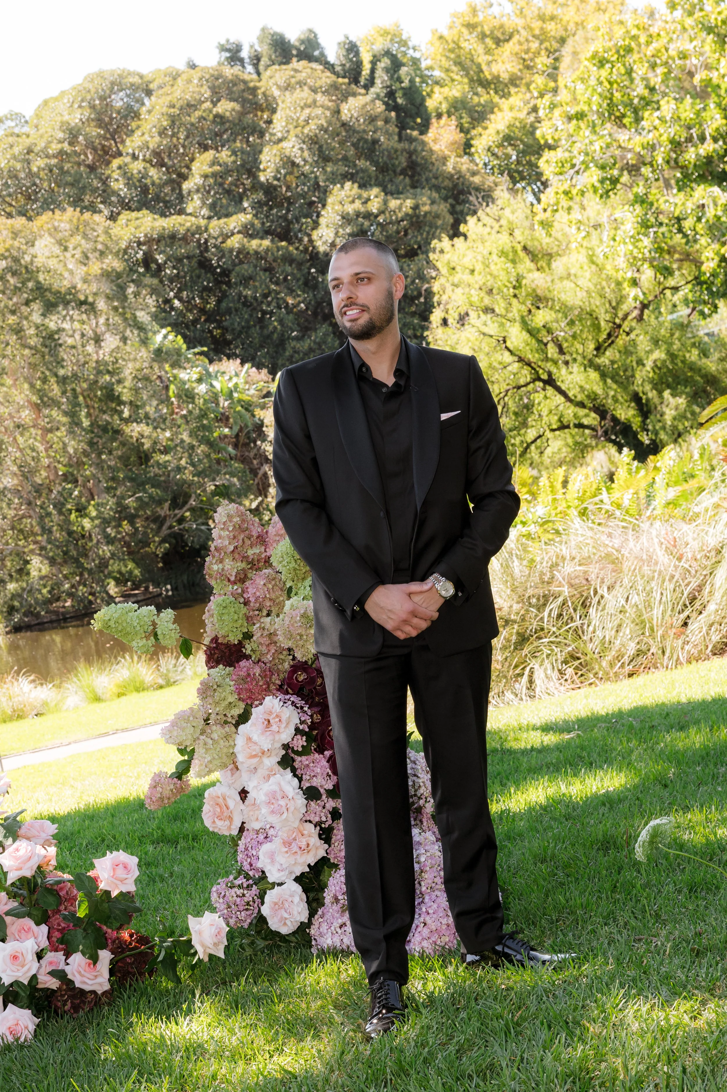 A man in a black suit and black dress shoes standing outdoors on grass, surrounded by pink and white flowers, with trees and a pond in the background during daytime.