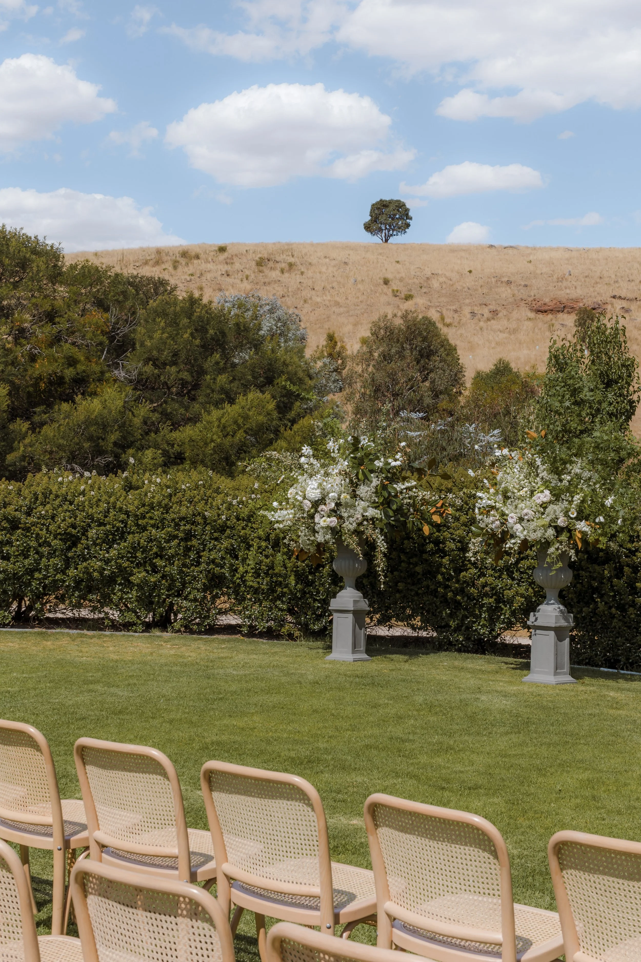 Outdoor wedding setup with empty beige chairs arranged on green grass, decorative flower arrangements in silver vases, against a backdrop of lush bushes, hills, a lone tree, and a partly cloudy sky.