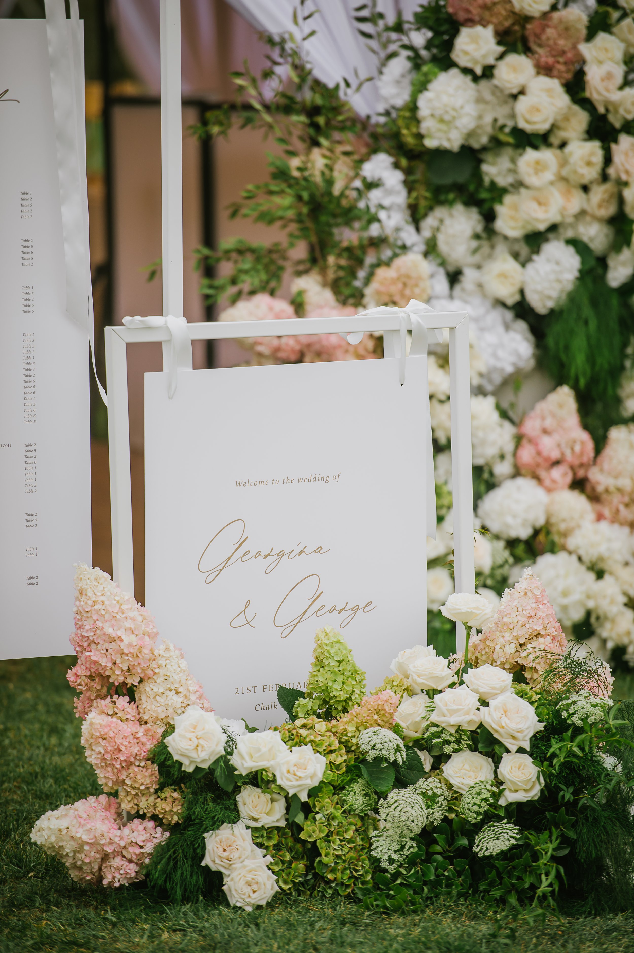 A wedding sign surrounded by a lush arrangement of white, pink, and cream flowers, with greenery, for Georgina and George's wedding on February 21st.