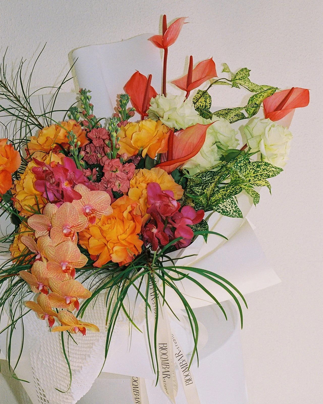 Colorful flower bouquet with pink, orange, yellow, and white flowers, including orchids, roses, and other blooms, arranged with green foliage, wrapped in white paper.