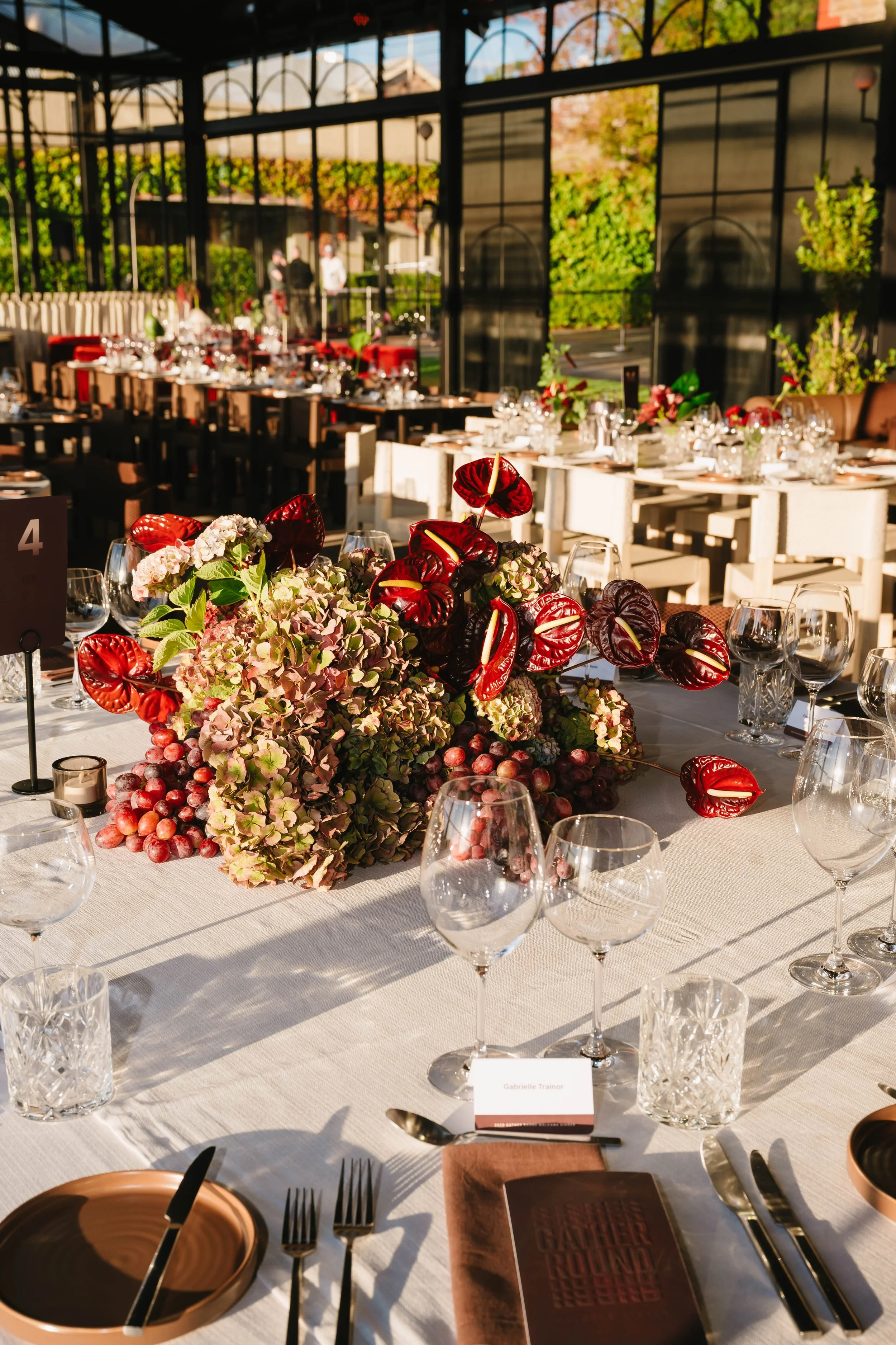 A decorated banquet table with a floral centerpiece, wine glasses, and place settings in a sunlit room with glass walls and a garden view.