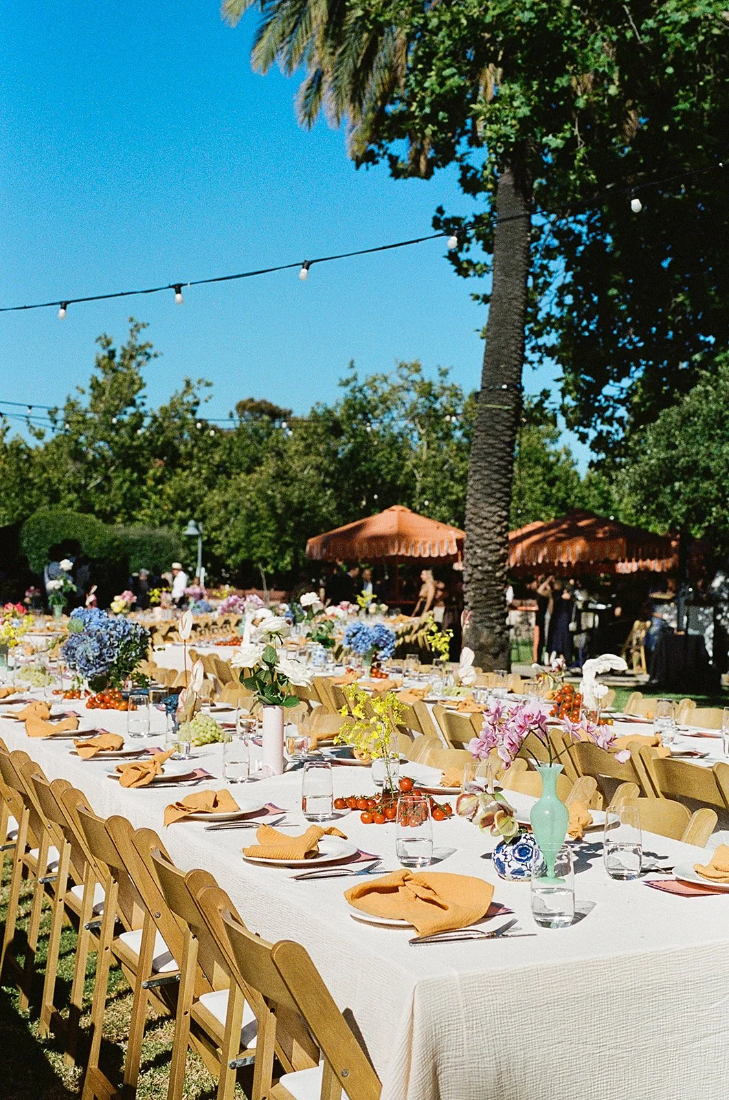 Outdoor banquet setup with long tables decorated with flowers, napkins, and glasses, under string lights and palm trees, on a sunny day.