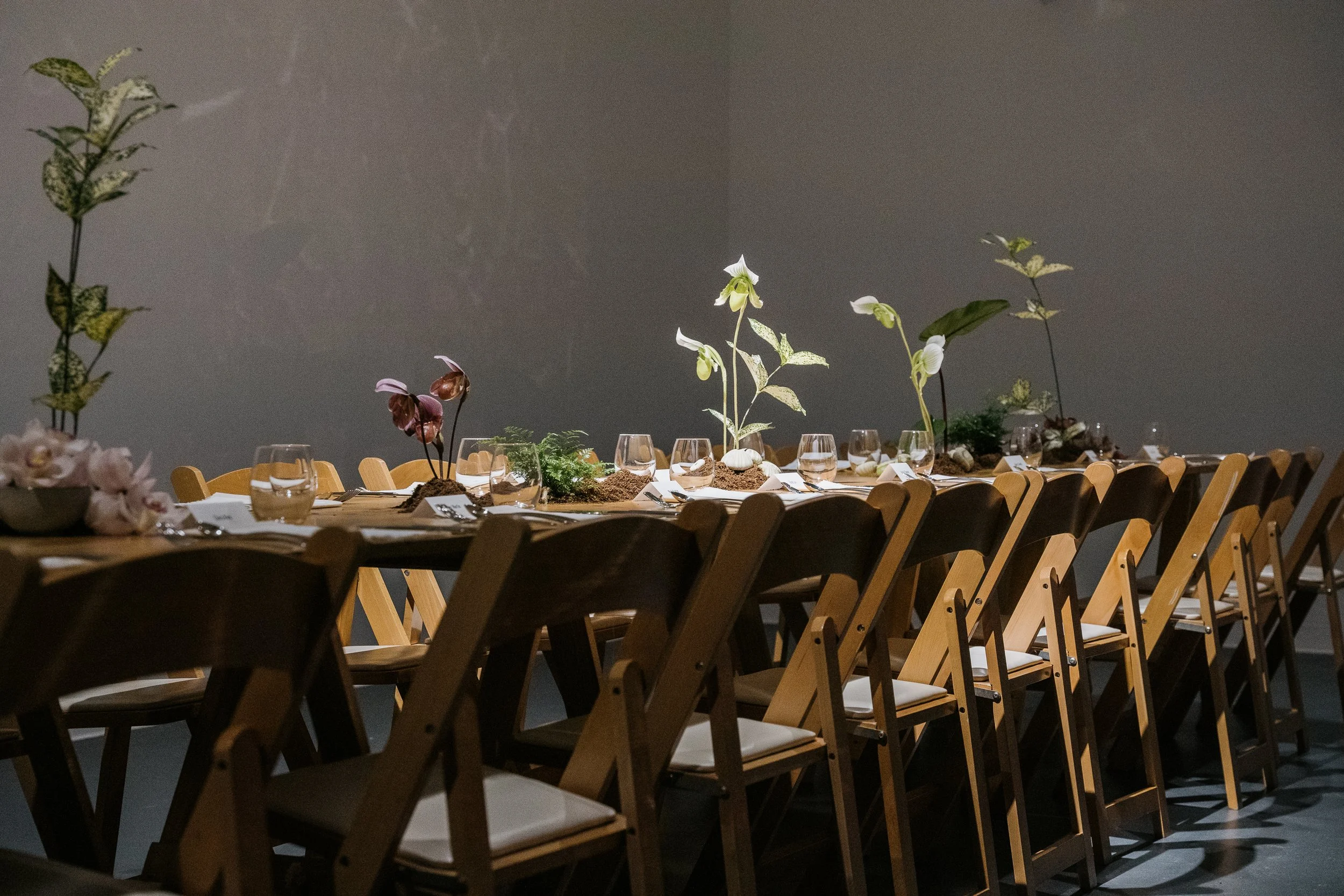 Long dining table decorated with plants and flowers, decorated as a nature-inspired centerpiece with chairs along both sides, against a plain wall background.