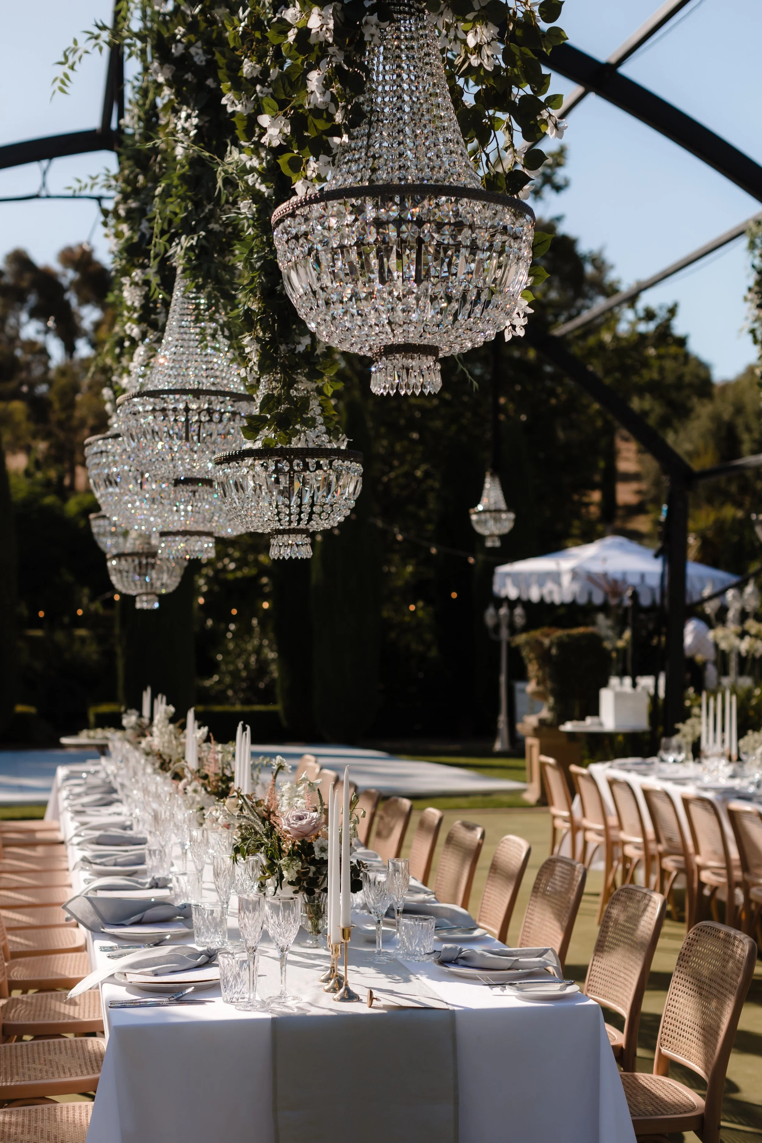Elegant outdoor event setup featuring a long dining table with white tablecloths, surrounded by chairs, and adorned with tall candlesticks and floral arrangements. Crystal chandeliers hang from a structure above the table, with trees and a white tent