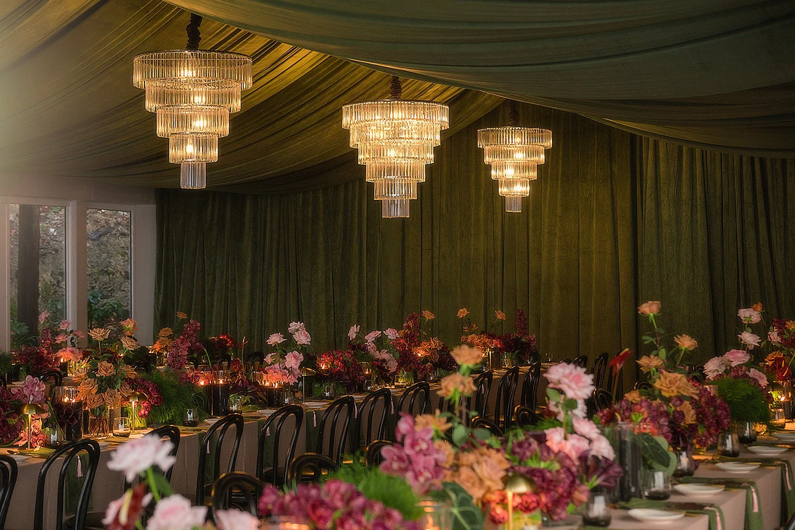 Elegant event space with a long dining table decorated with pink, red, and purple flowers, surrounded by black chairs. The ceiling is draped with fabric, and there are three large crystal chandeliers hanging overhead, casting a warm glow. In the back