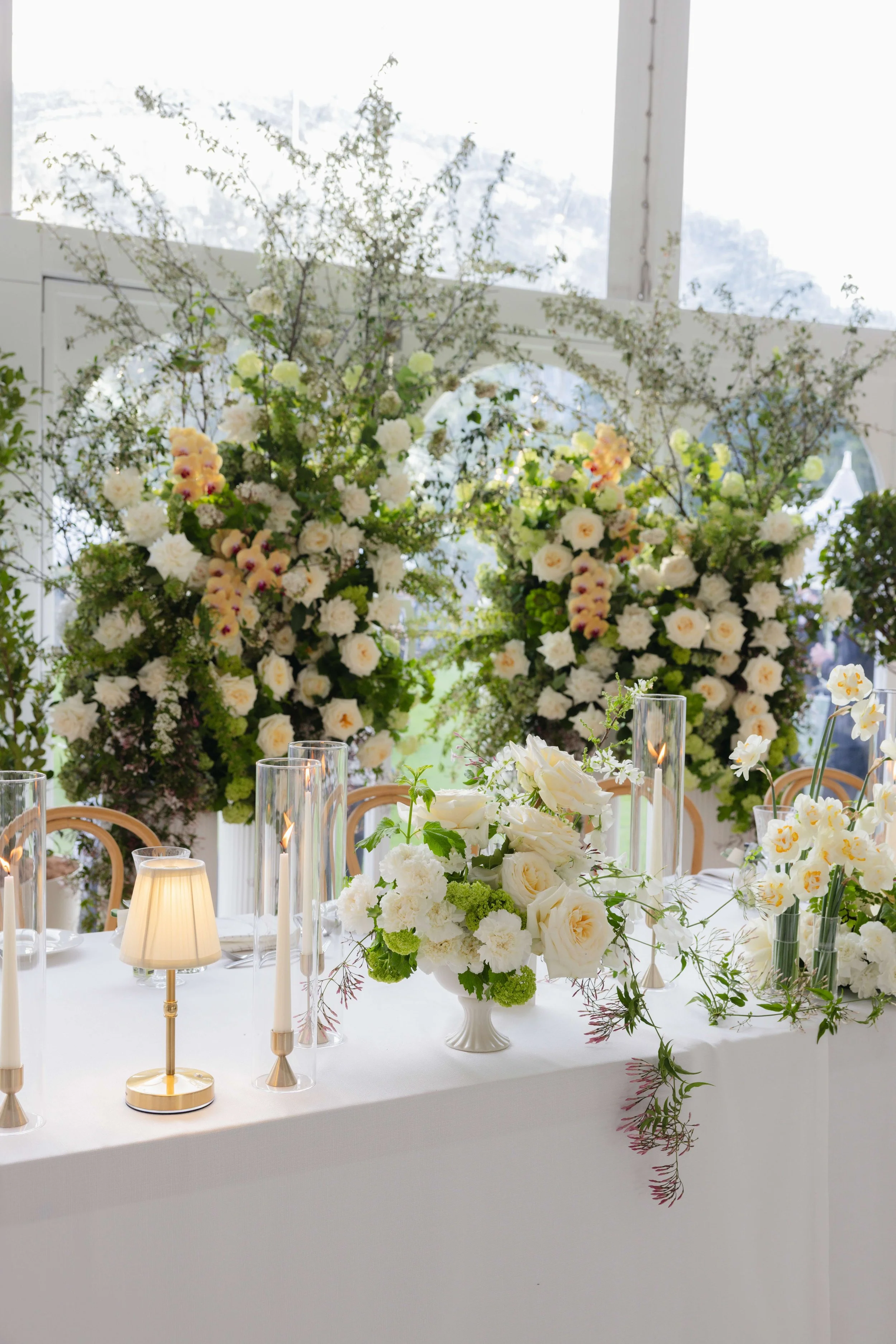 Elegant event table with white flowers, tall glass candle holders, and small lamps, set against a large floral arrangement of white and pink flowers and lush greenery.