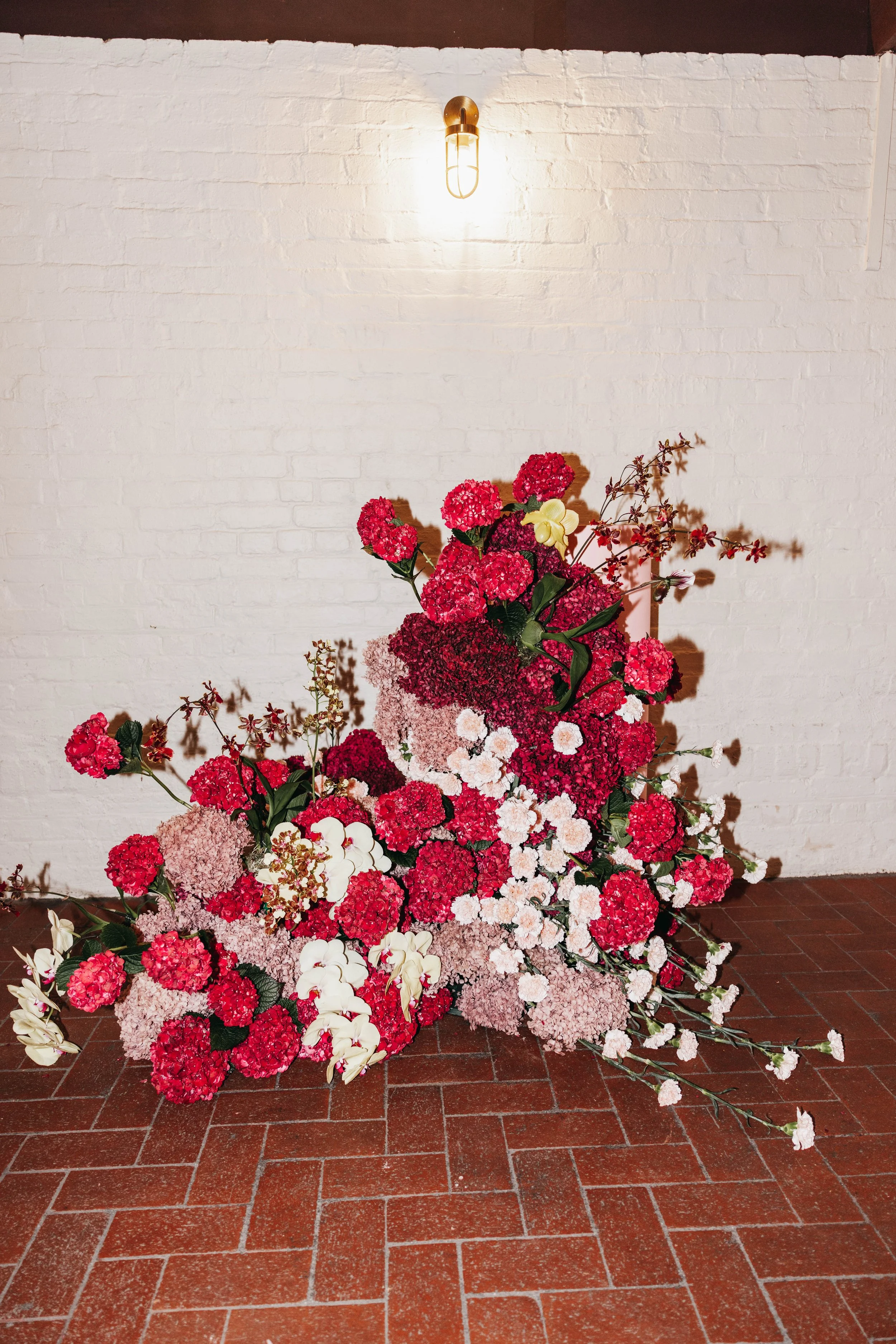A large floral arrangement with pink, white, and red flowers, cascading onto a brick floor against a white brick wall, illuminated by a wall-mounted light fixture.