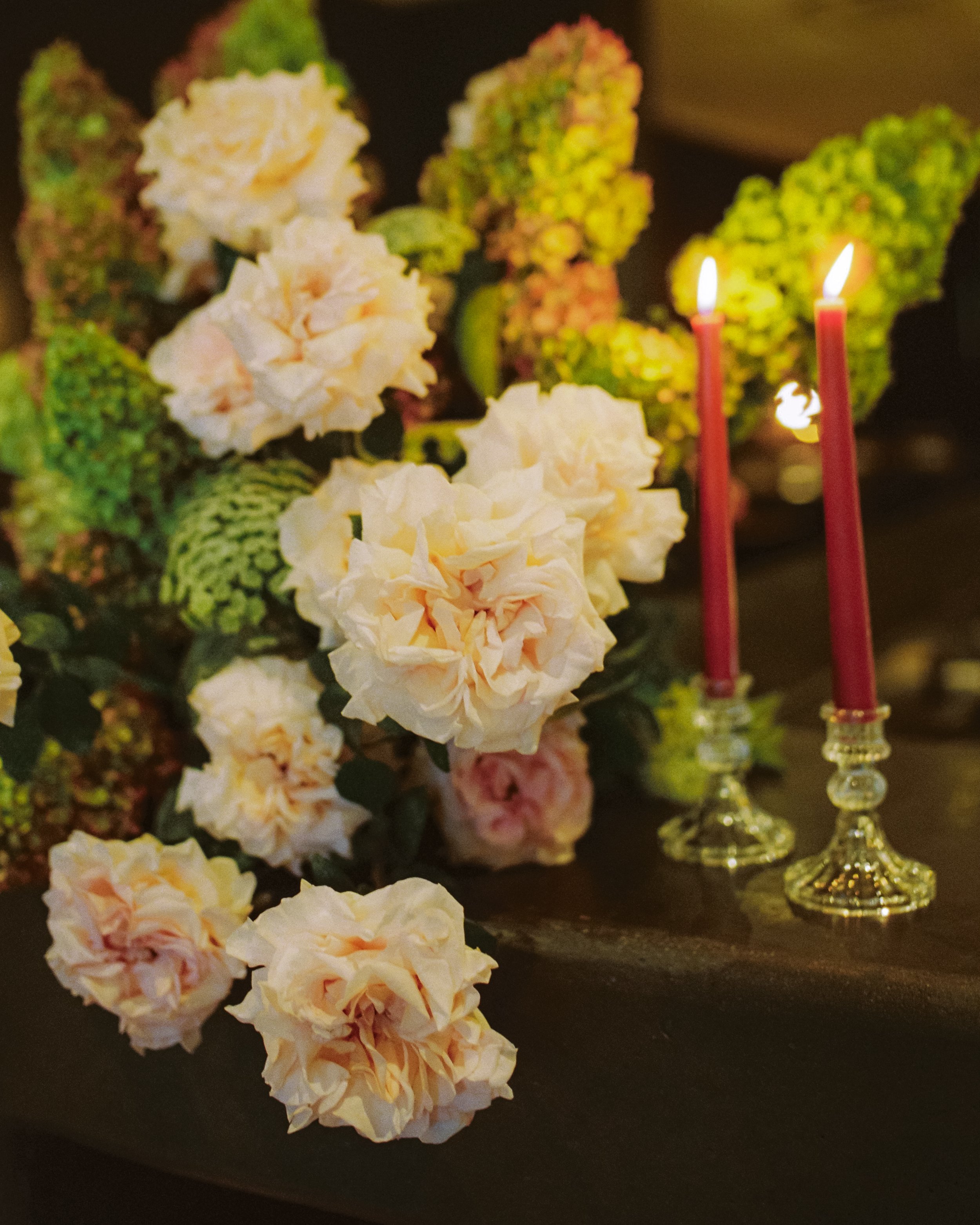 Cream-colored artificial flowers and red candles in gold holders on a black surface.