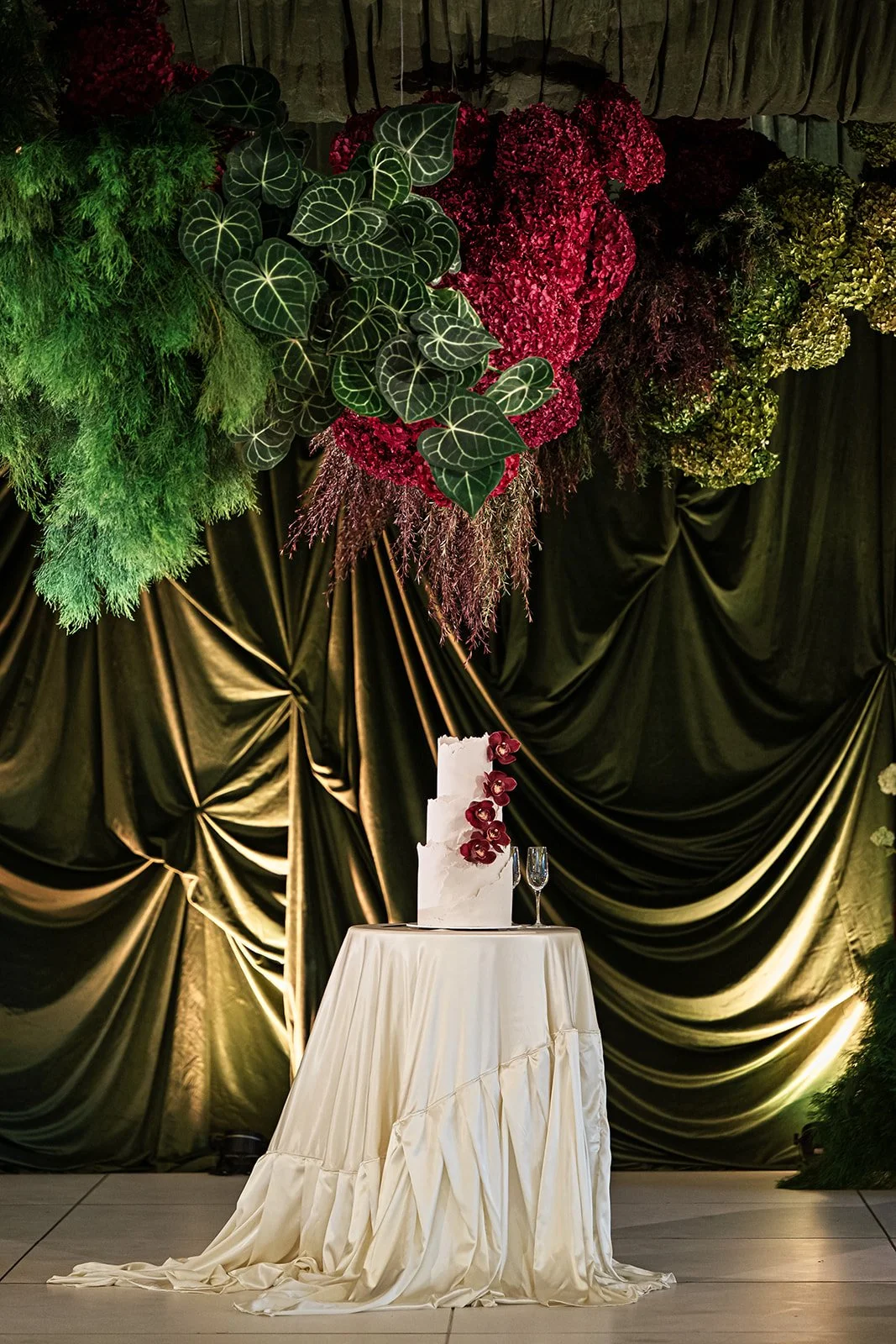 A wedding cake with orchids on a table in front of a backdrop of black curtain and large floral arrangement. The arrangement includes green leaves, red and pink flowers, and draped greenery.