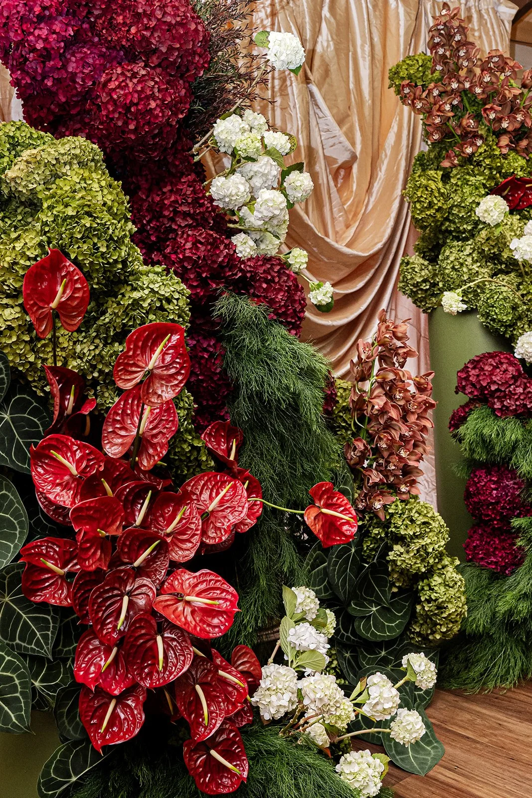 Arrangement of various colorful flowers, including red anthuriums, green and white hydrangeas, and pink orchids, with a gold fabric backdrop.
