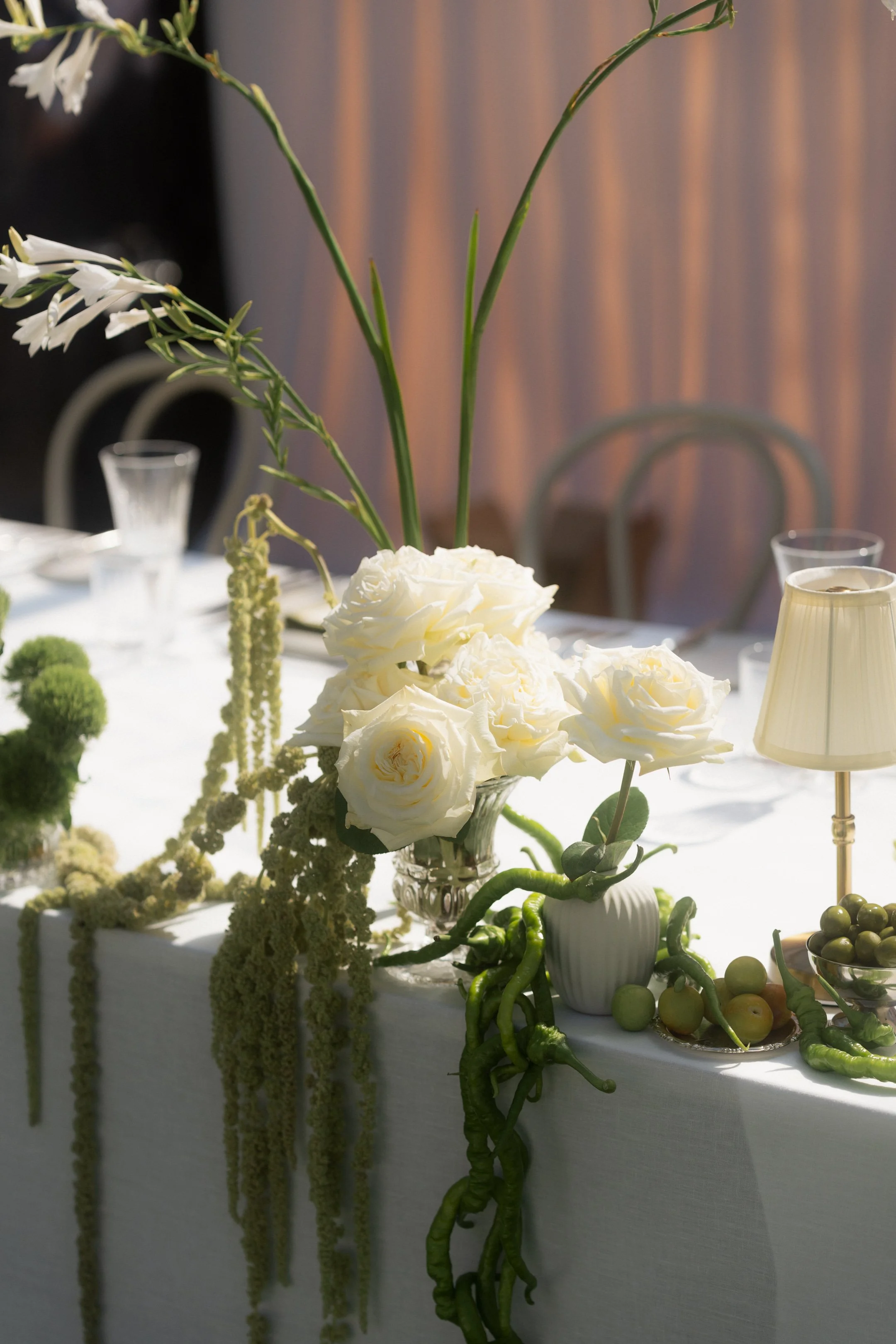 A floral arrangement with white roses and green foliage on a table, with additional white flowers and decorative elements in the background.