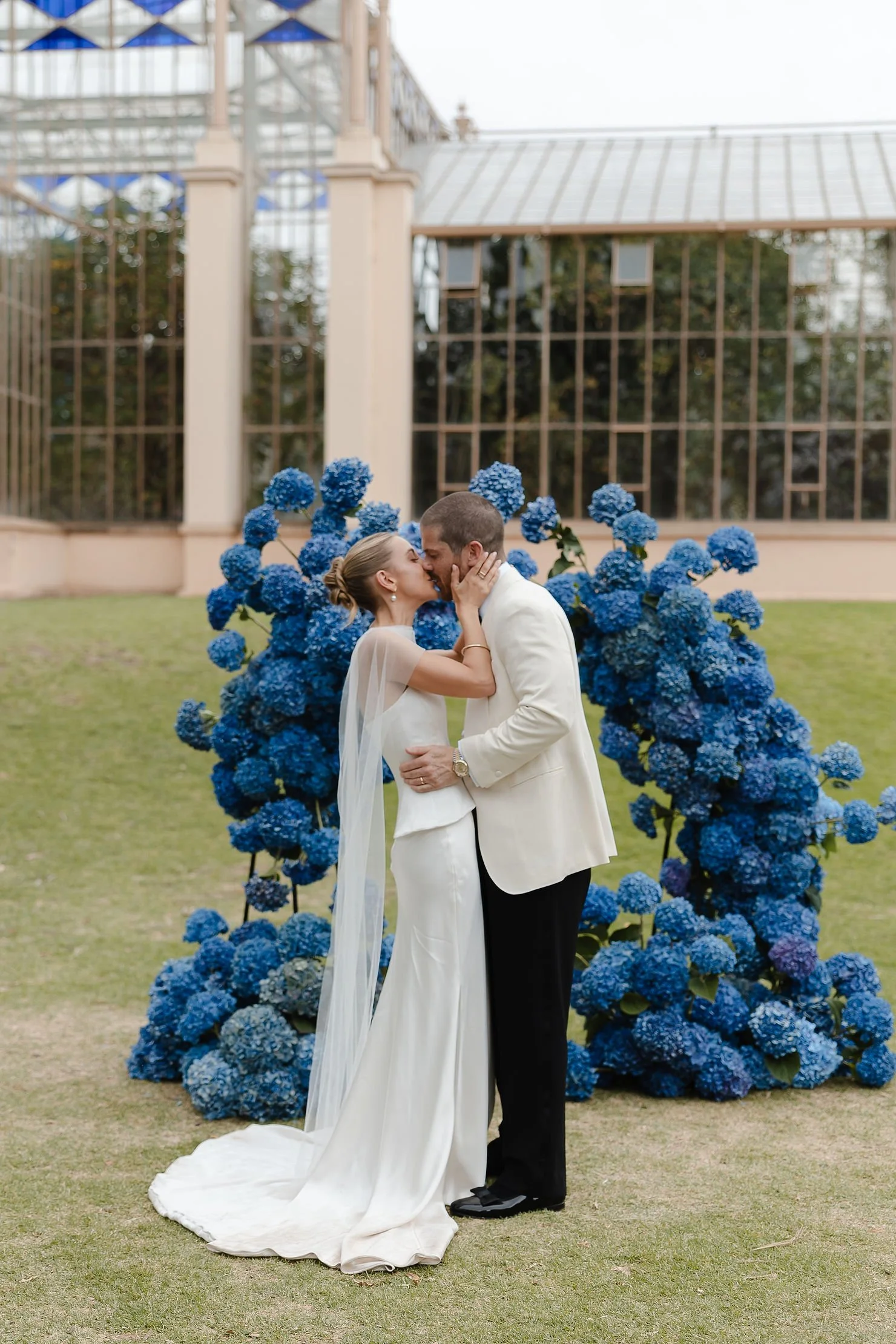 A wedding altar decorated with white flowers and greenery inside a glass-walled structure, with white lamps hanging from above.