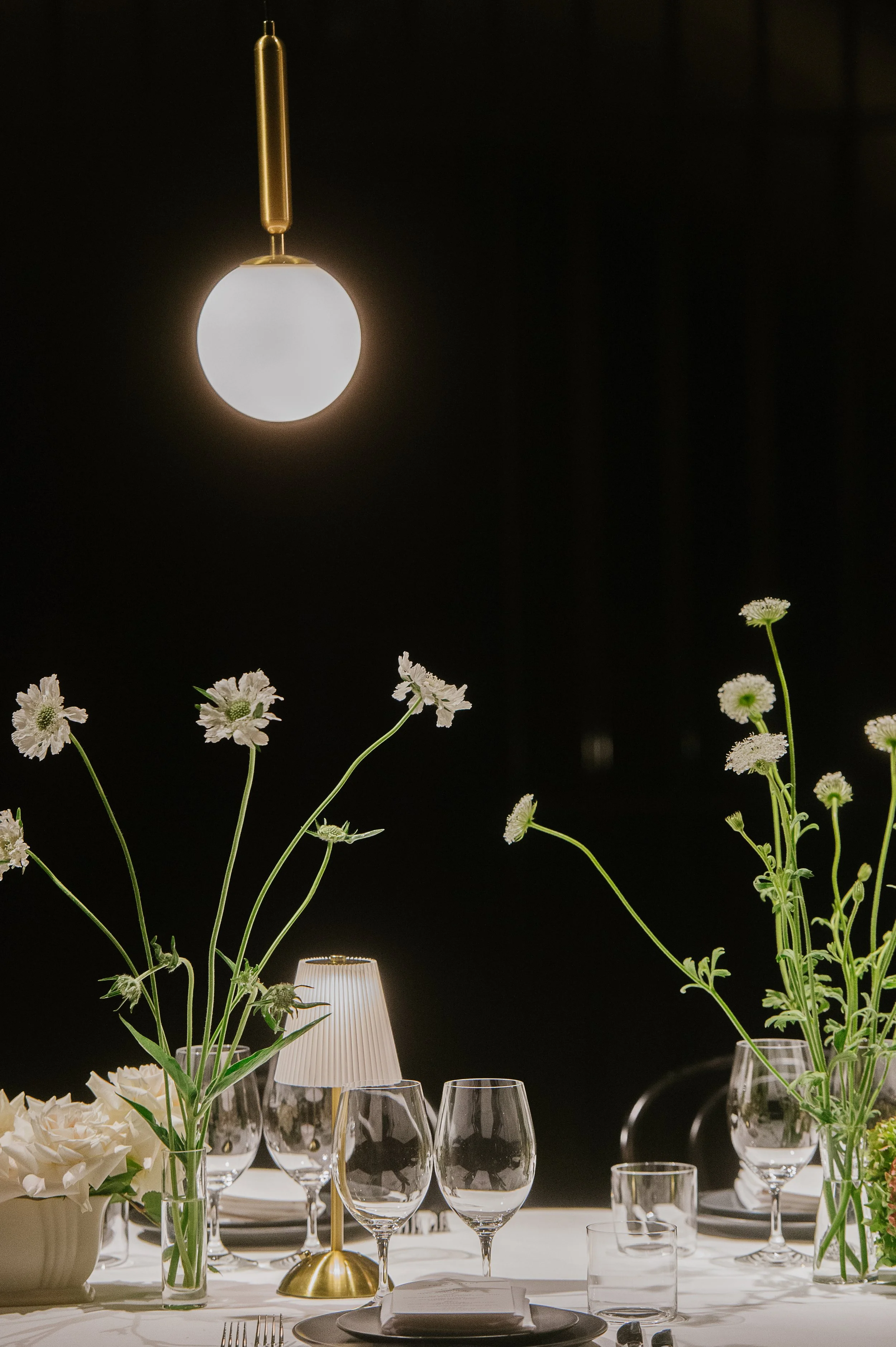 Elegant dining table set with white flowers, wine glasses, and a brass table lamp against a dark background.
