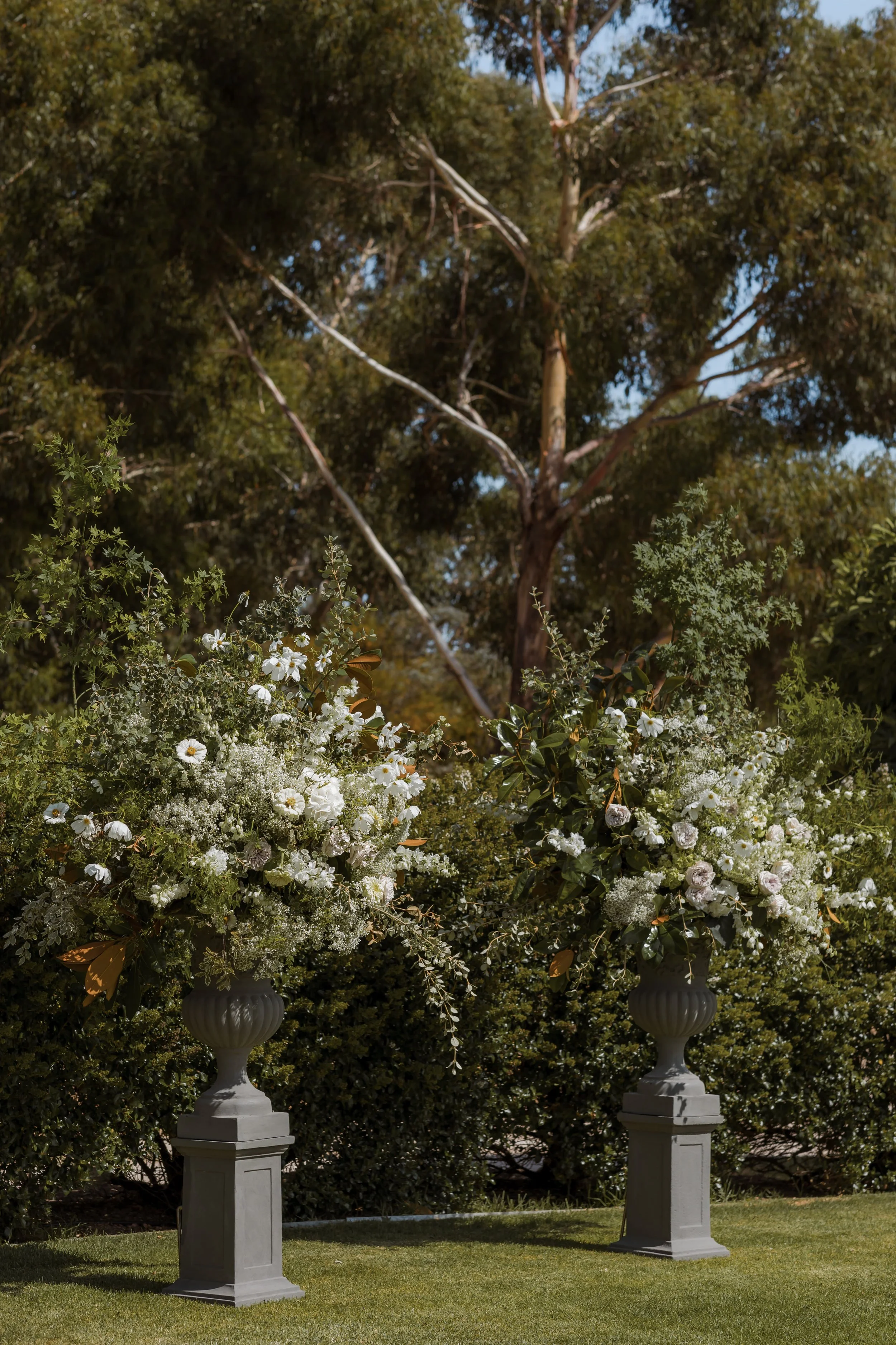 Two large floral arrangements in gray urn vases on pedestals, placed on a grassy lawn against a backdrop of dense trees and foliage.