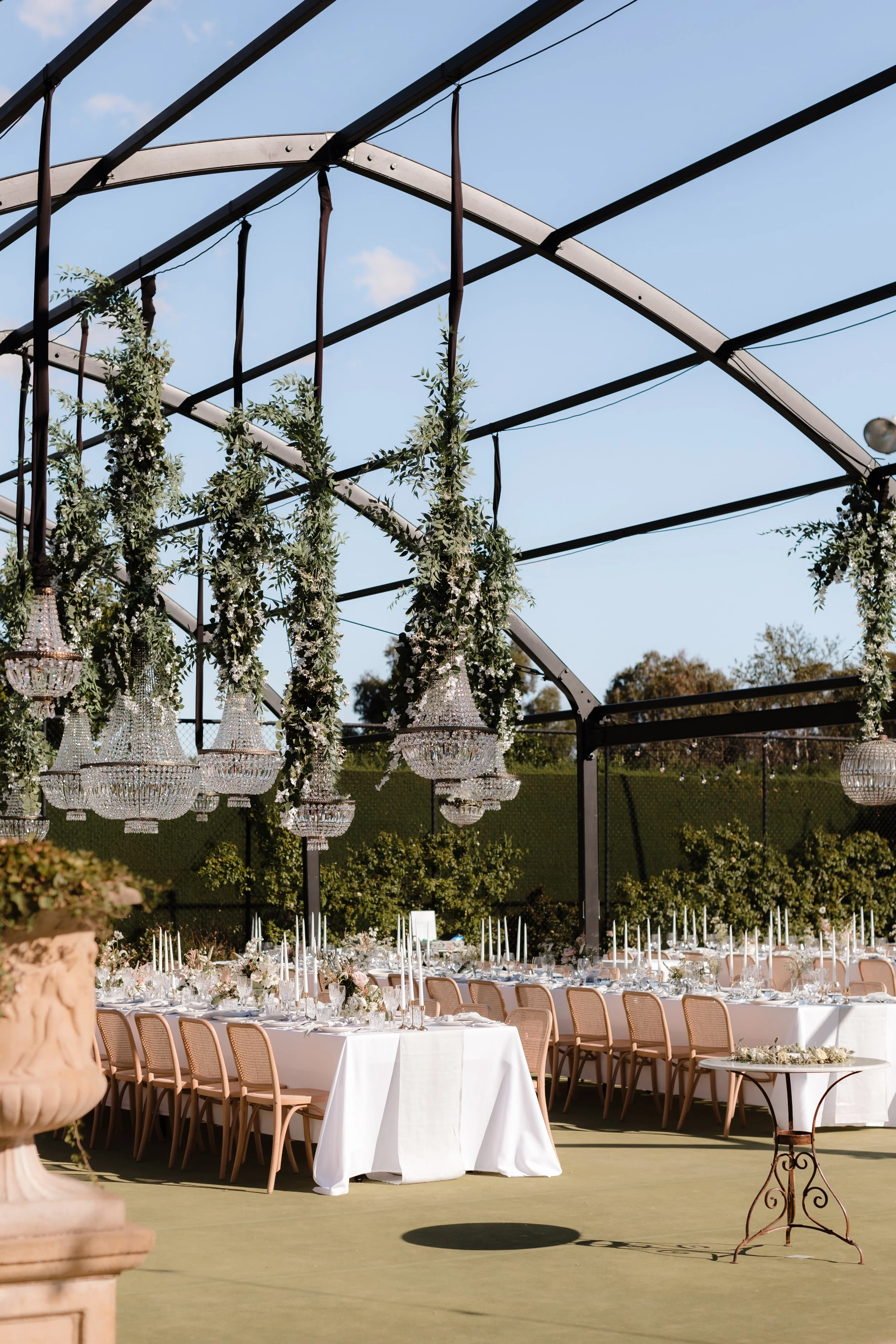 Outdoor event setup with long dining tables, white tablecloths, floral centerpieces, tall candlesticks, and hanging chandeliers under a black metal framework with greenery and a blue sky.
