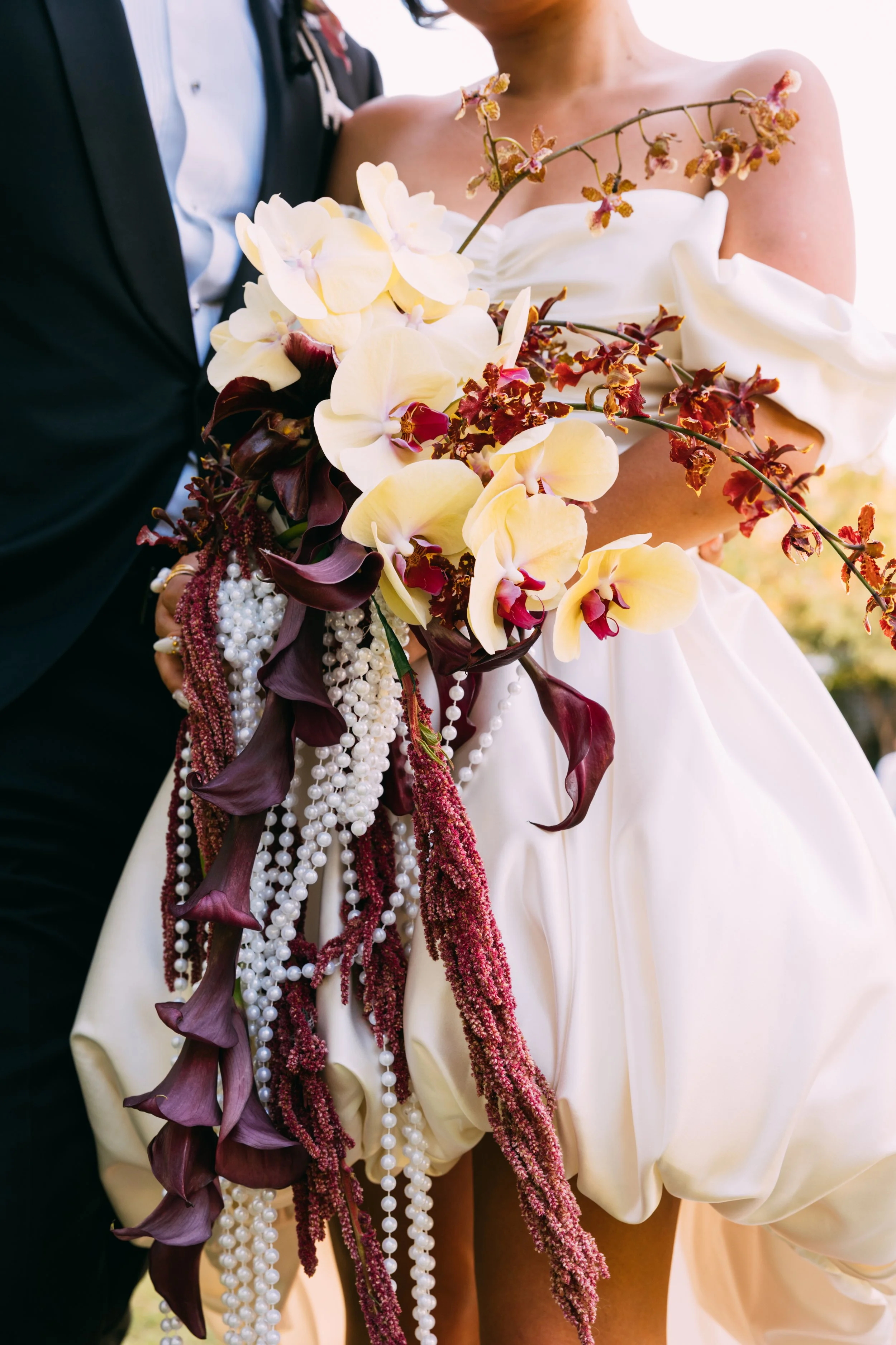 A bride wearing a white dress holding a bouquet of large white orchids, purple Calla lilies, and hanging pearl and burgundy flower strands, with a groom in a black tuxedo beside her.