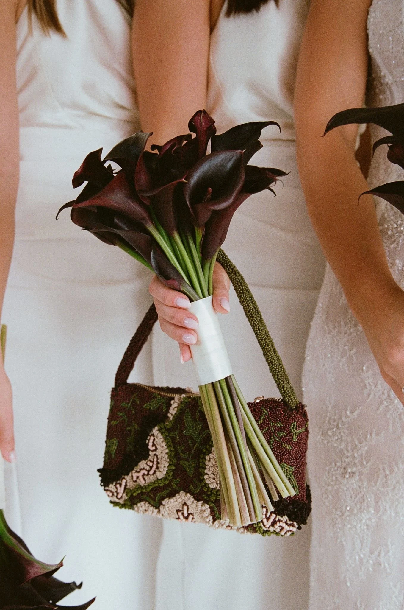 A person holding a bouquet of dark purple calla lilies and wearing a patterned skirt with a white dress and another person in an off-white dress standing nearby.