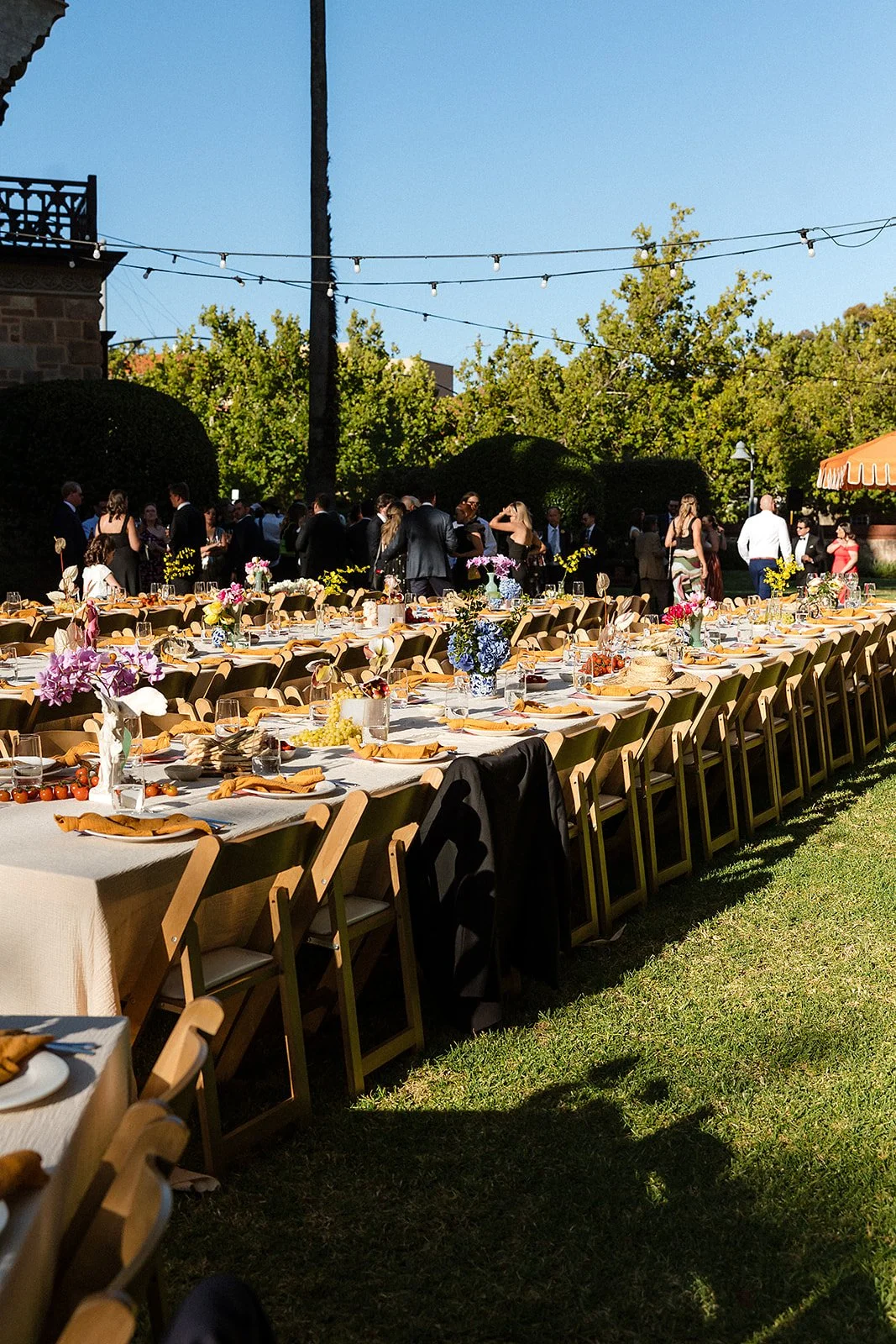 Outdoor banquet setup with long tables decorated with flowers, set for a formal event with the backdrop of trees and guests mingling.