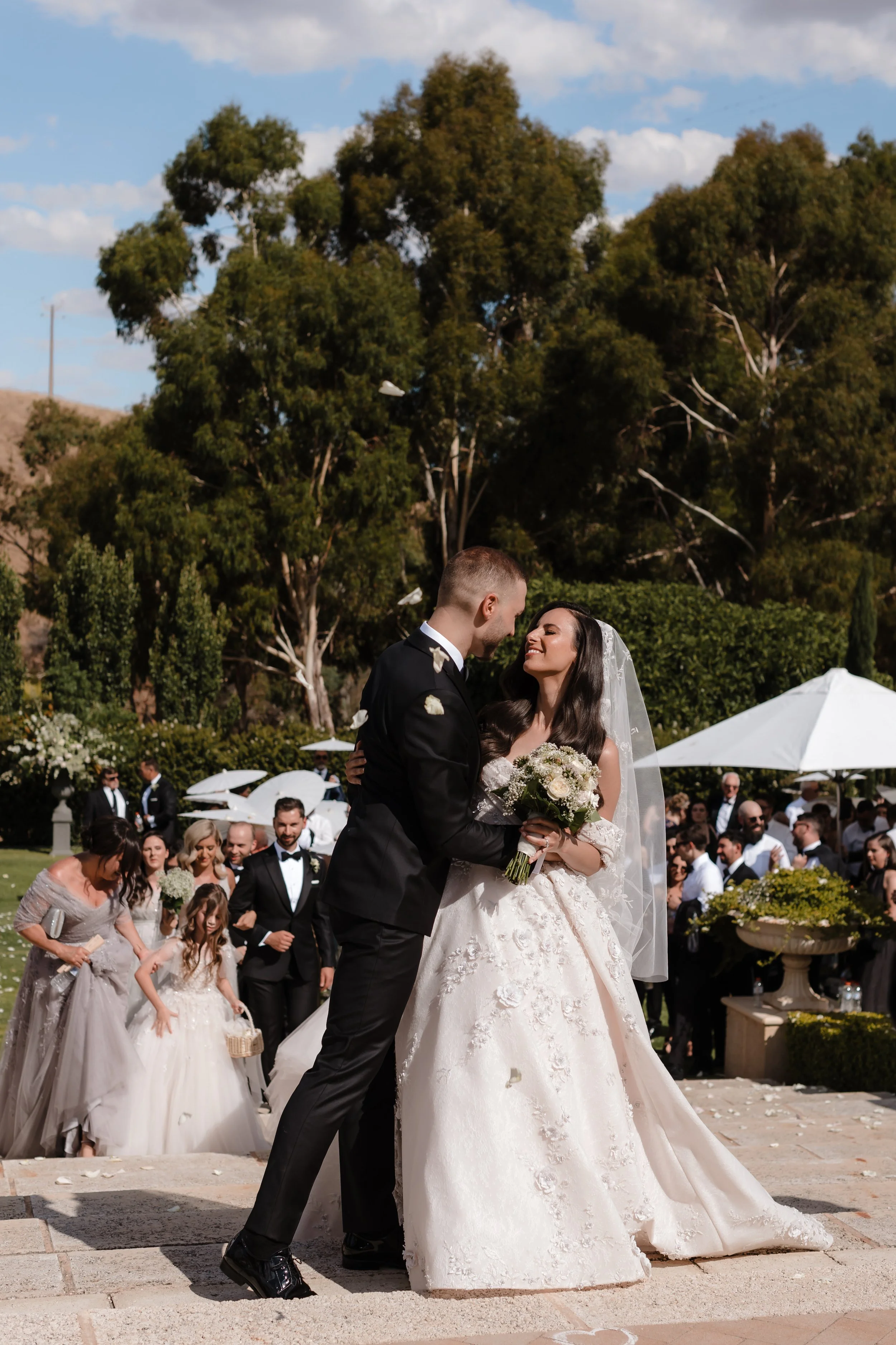 A wedding ceremony outdoors with a bride and groom sharing a moment, surrounded by guests and trees in the background.