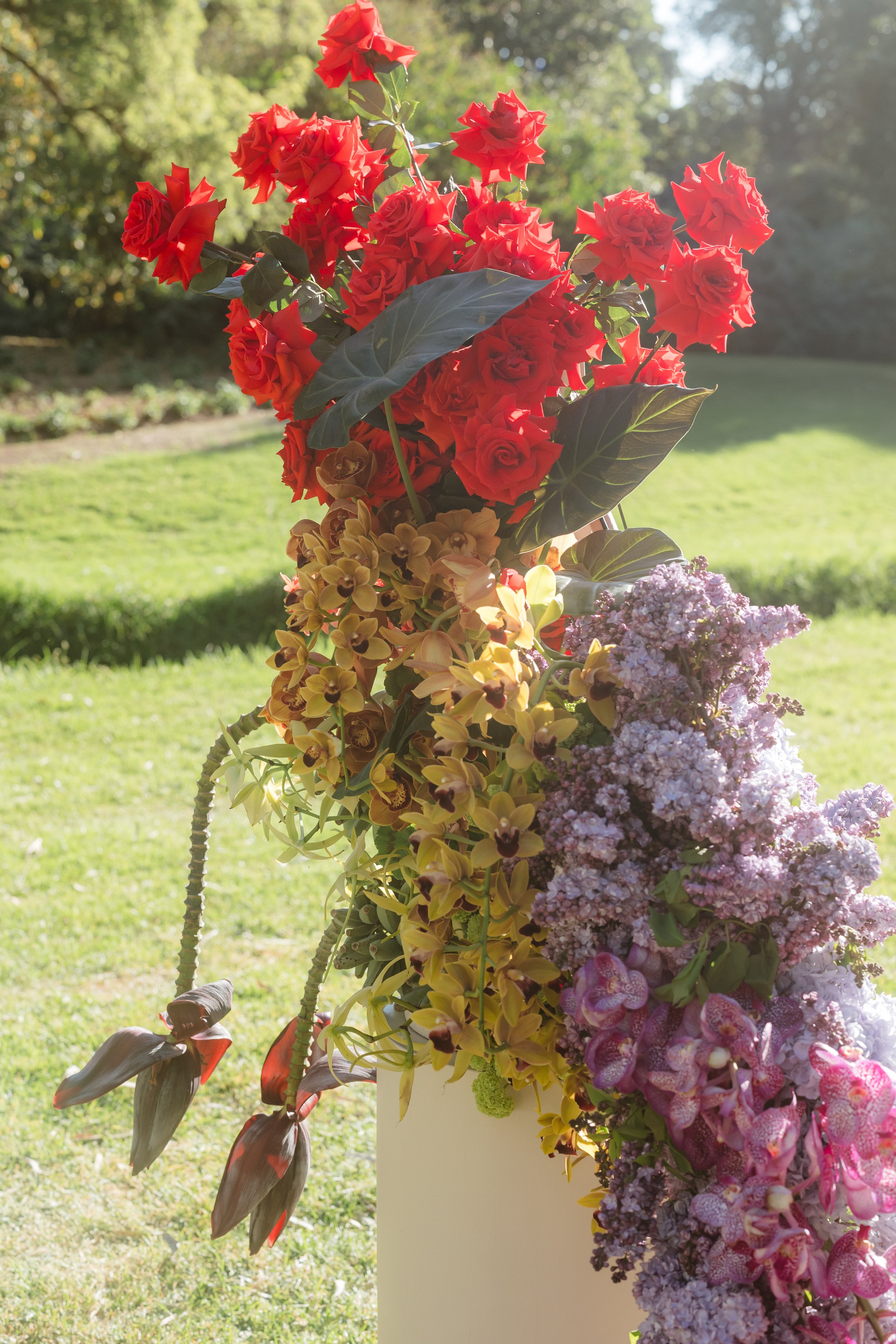 An arrangement of various colorful flowers, including red roses, purple hydrangeas, and yellow orchids, displayed outdoors in a garden setting with lush greenery in the background.