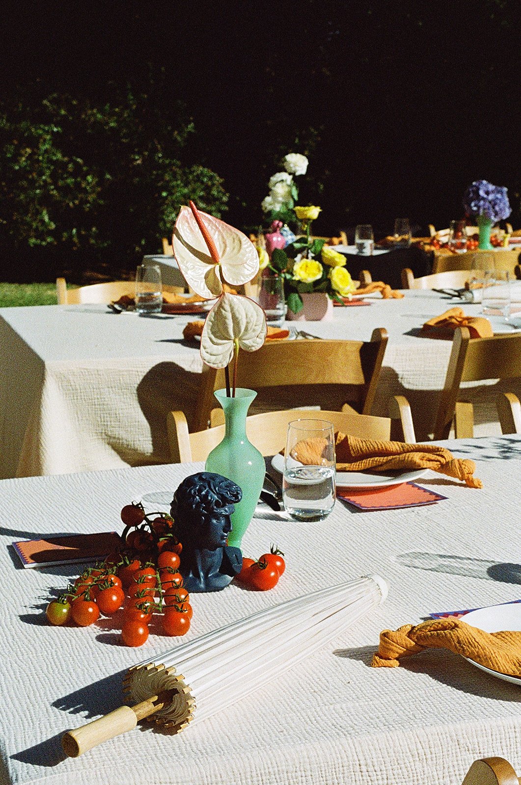 Outdoor dining table decorated with a vase holding folded cloth flowers, cherry tomatoes, a small black sculpture of a head, a fan, water glass, and napkins, set in a garden at night.