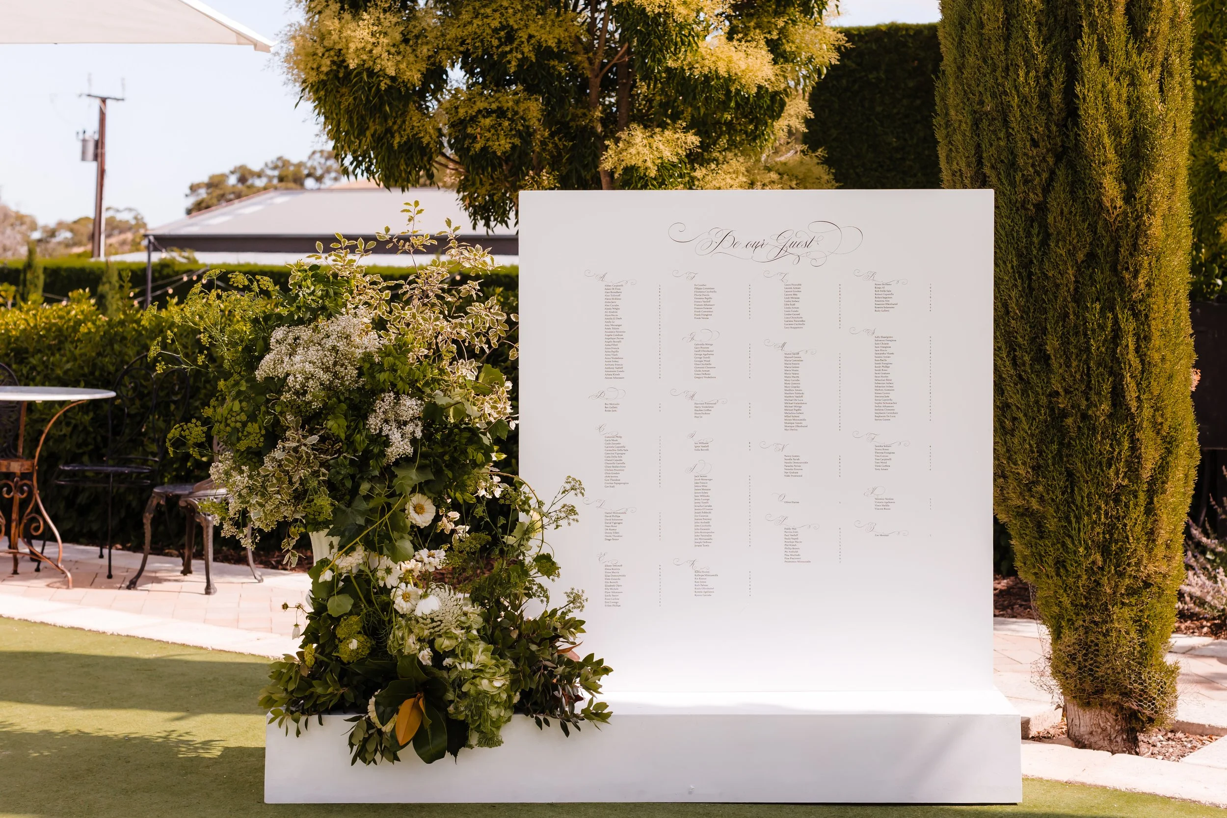 Outdoor wedding seating chart on a white board decorated with flowers and greenery, outdoors with trees and bushes in the background.