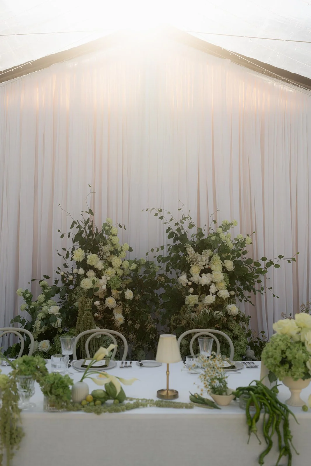Elegant wedding or event table setup with floral arrangements, white draped backdrop, and ambient lighting.