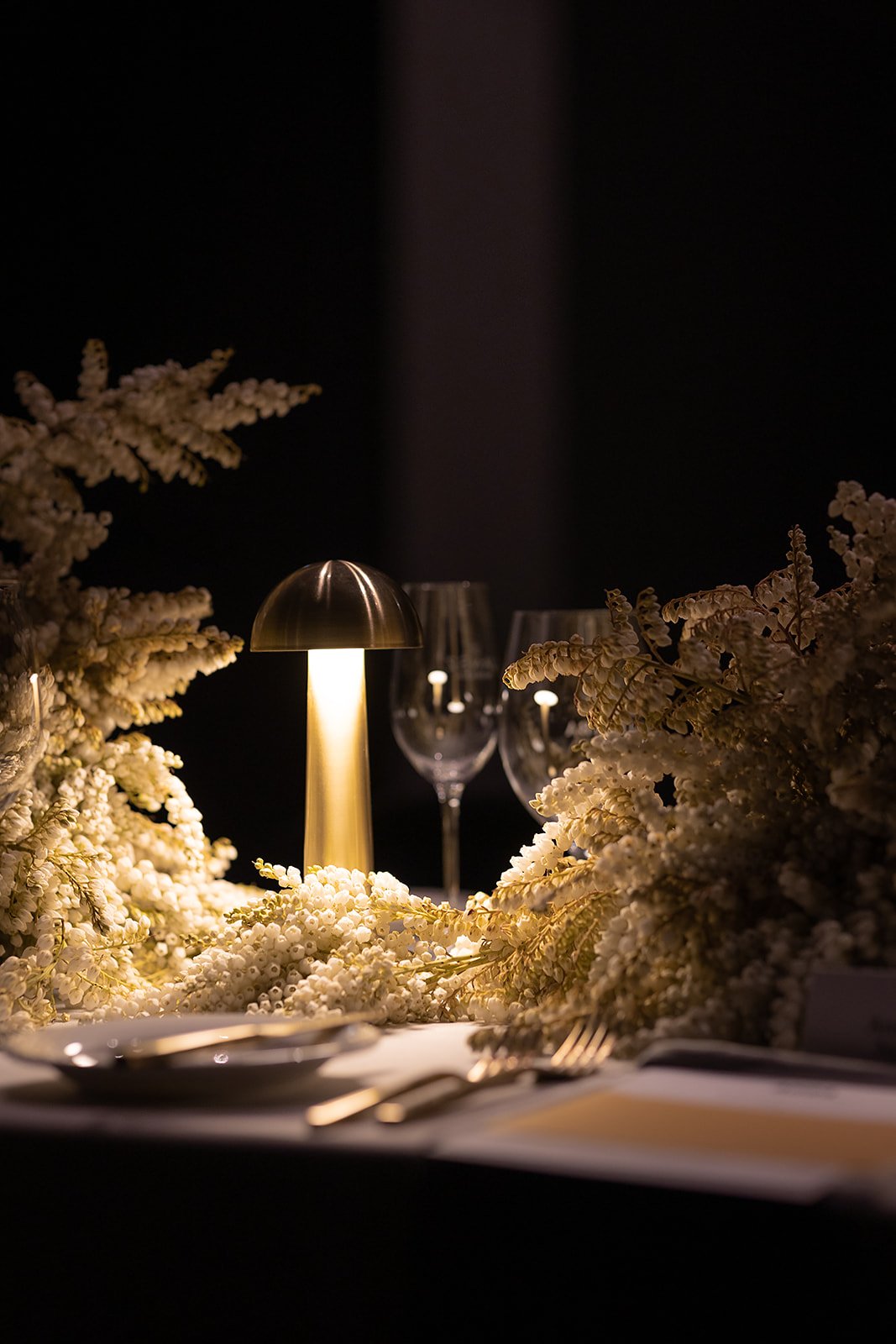 Elegant dining table with white floral arrangements, a modern table lamp, and wine glasses, illuminated in a dark setting.