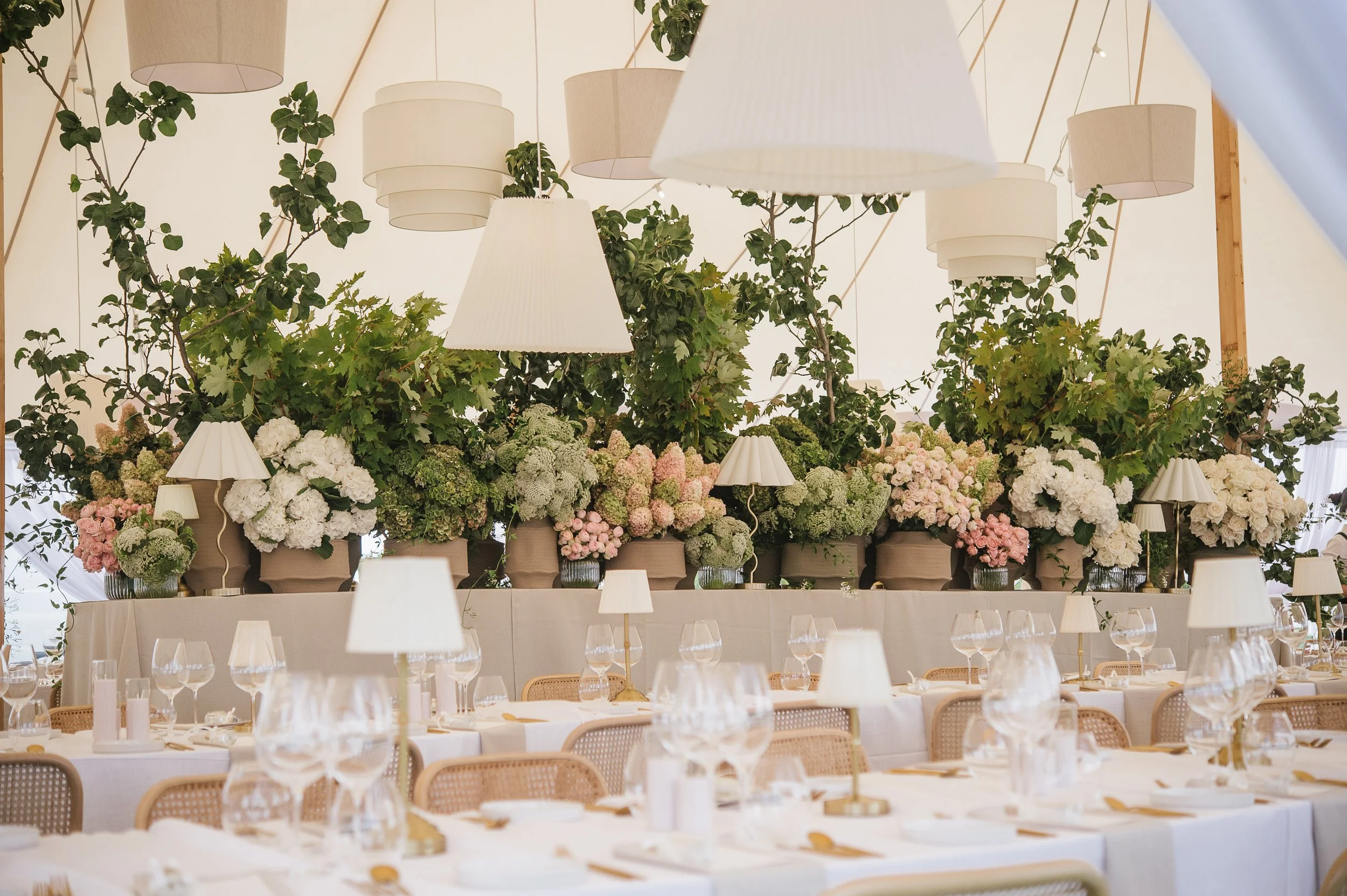 Elegant indoor event space decorated with numerous potted plants and flowers, with hanging pendant lights and set tables with glasses and napkins.
