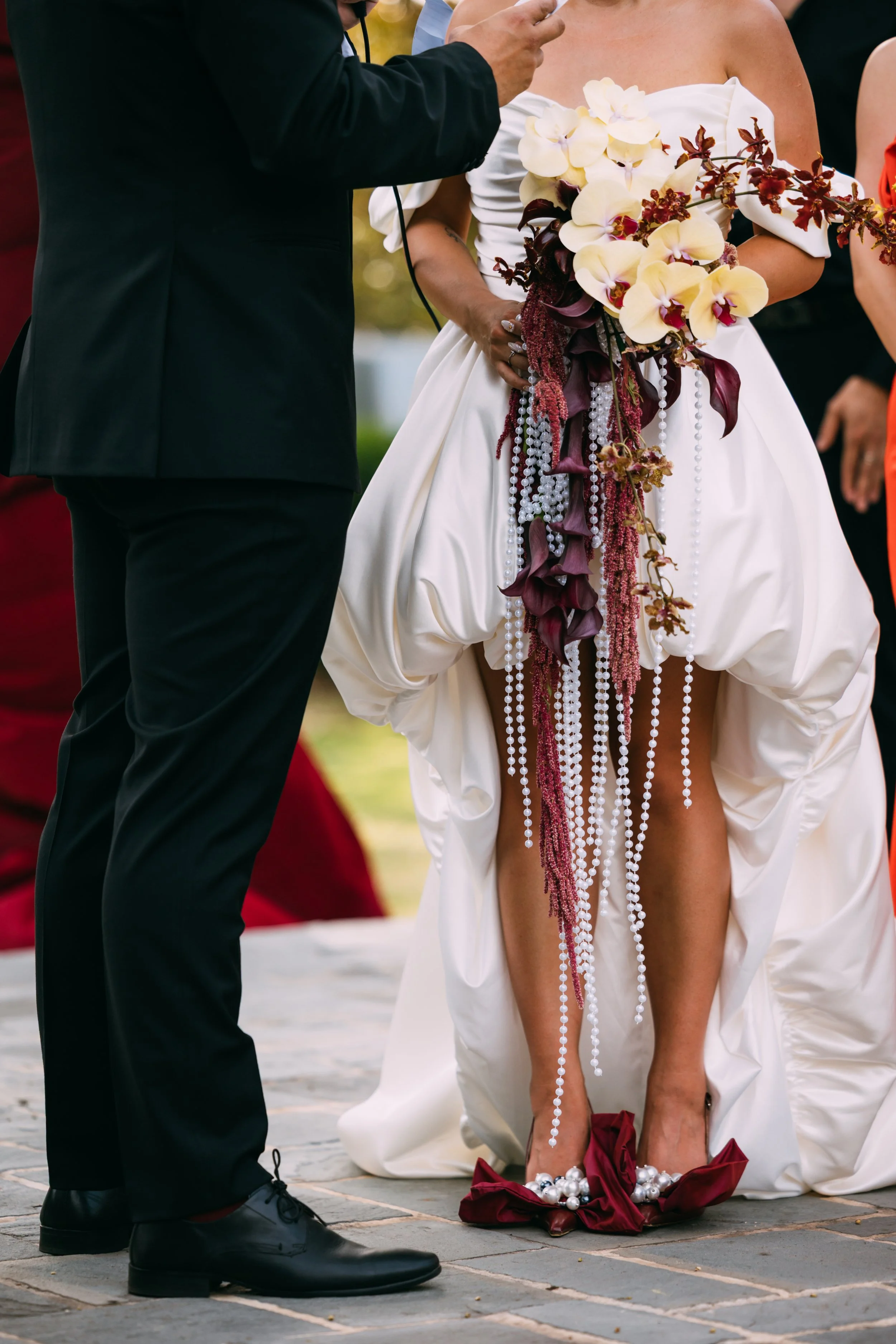 A woman in a wedding dress kneels on the ground, holding a bouquet of cream and burgundy flowers. She is wearing red shoes with pearl embellishments and a white dress with a gathered front. A man in a black suit and shoes stands nearby, with his hand extended towards her, possibly during a wedding ceremony.