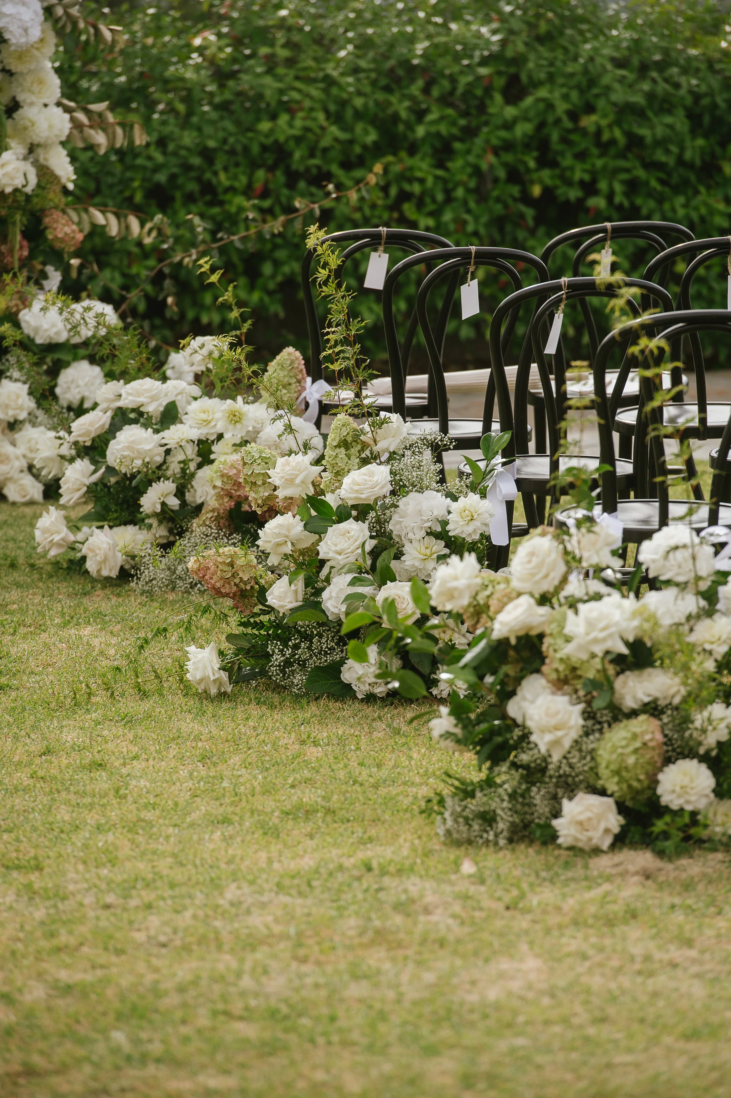 Black chairs with white tags arranged in a row on green grass, decorated with white and green flowers for a wedding or outdoor event.