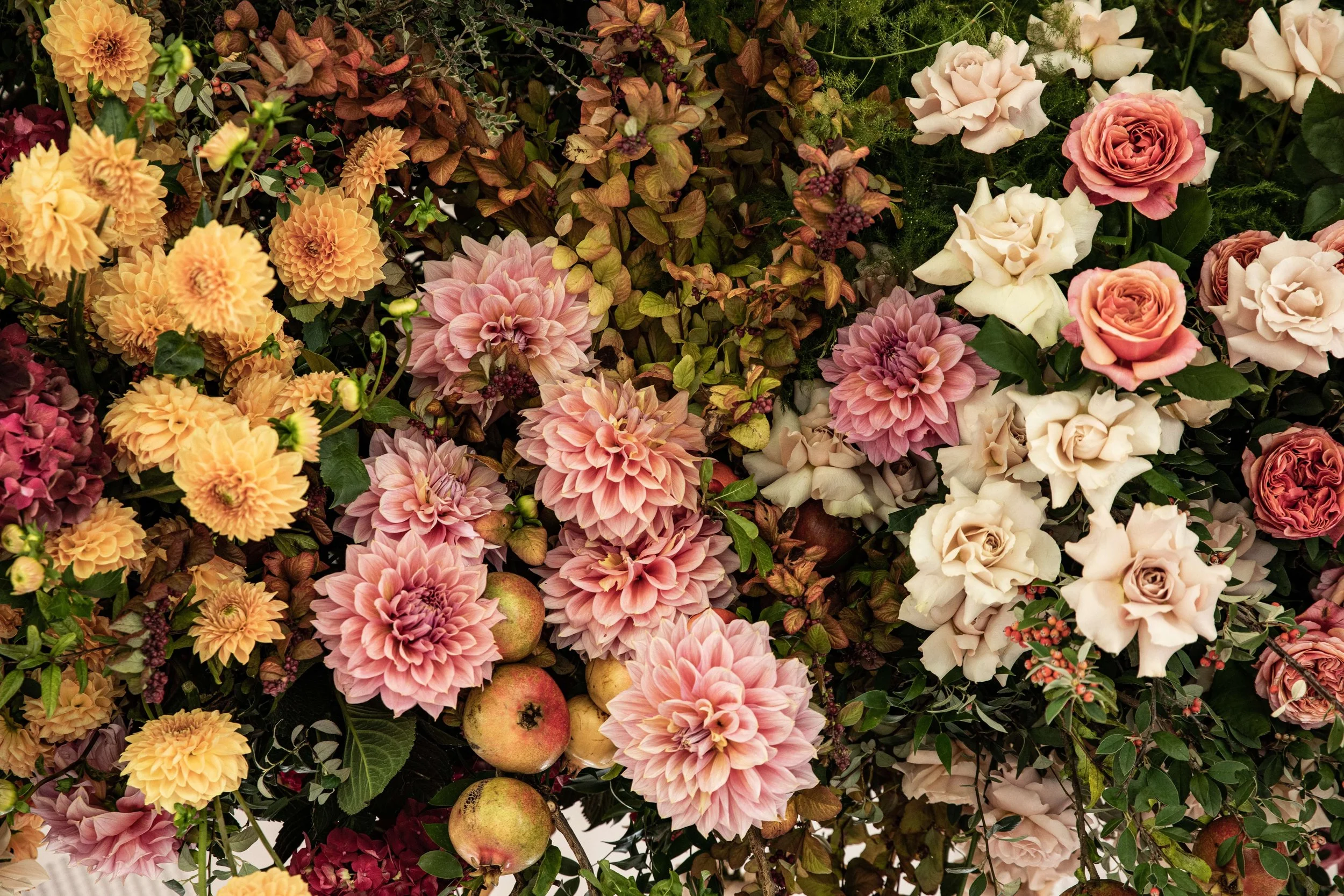 A large bouquet of various flowers including dahlias, roses, and other blossoms in shades of peach, pink, white, and purple, with some small apples among the flowers and green foliage.