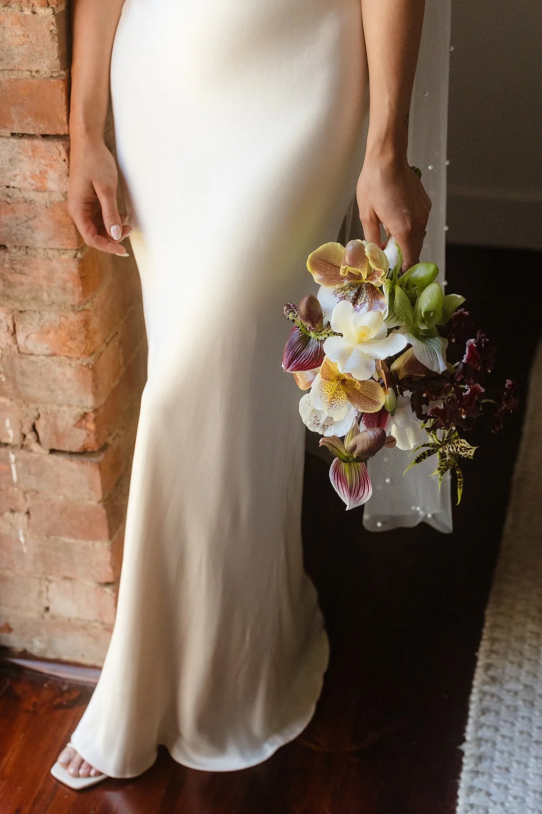 Person in a white satin dress holding a bouquet of mixed flowers, standing on a wooden floor next to a brick wall.