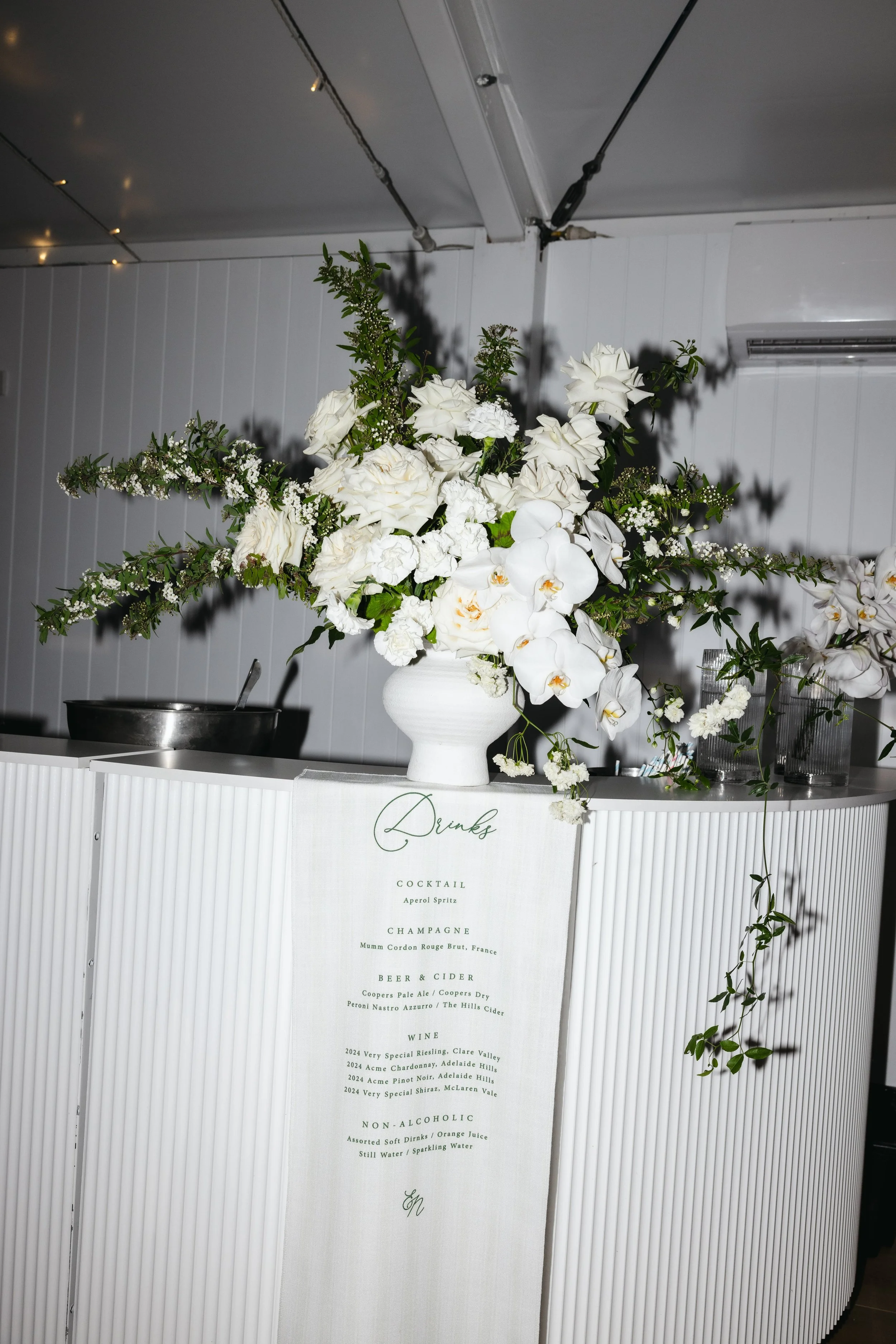 A large white floral arrangement with white roses, orchids, and greenery in a white vase on a table. Behind the arrangement, a menu with drink, wine, and non-alcoholic options is visible. The background features a white paneled wall and ceiling.