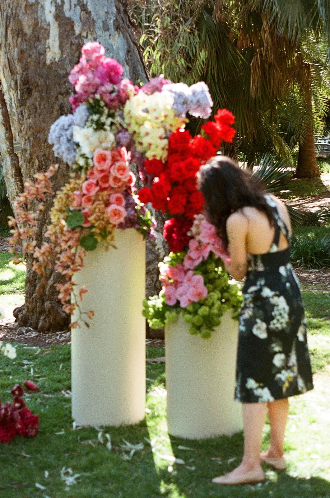 A woman with long dark hair, wearing a black dress with white floral patterns, is looking at a floral arrangement on white pedestals outdoors in a park with trees and greenery.