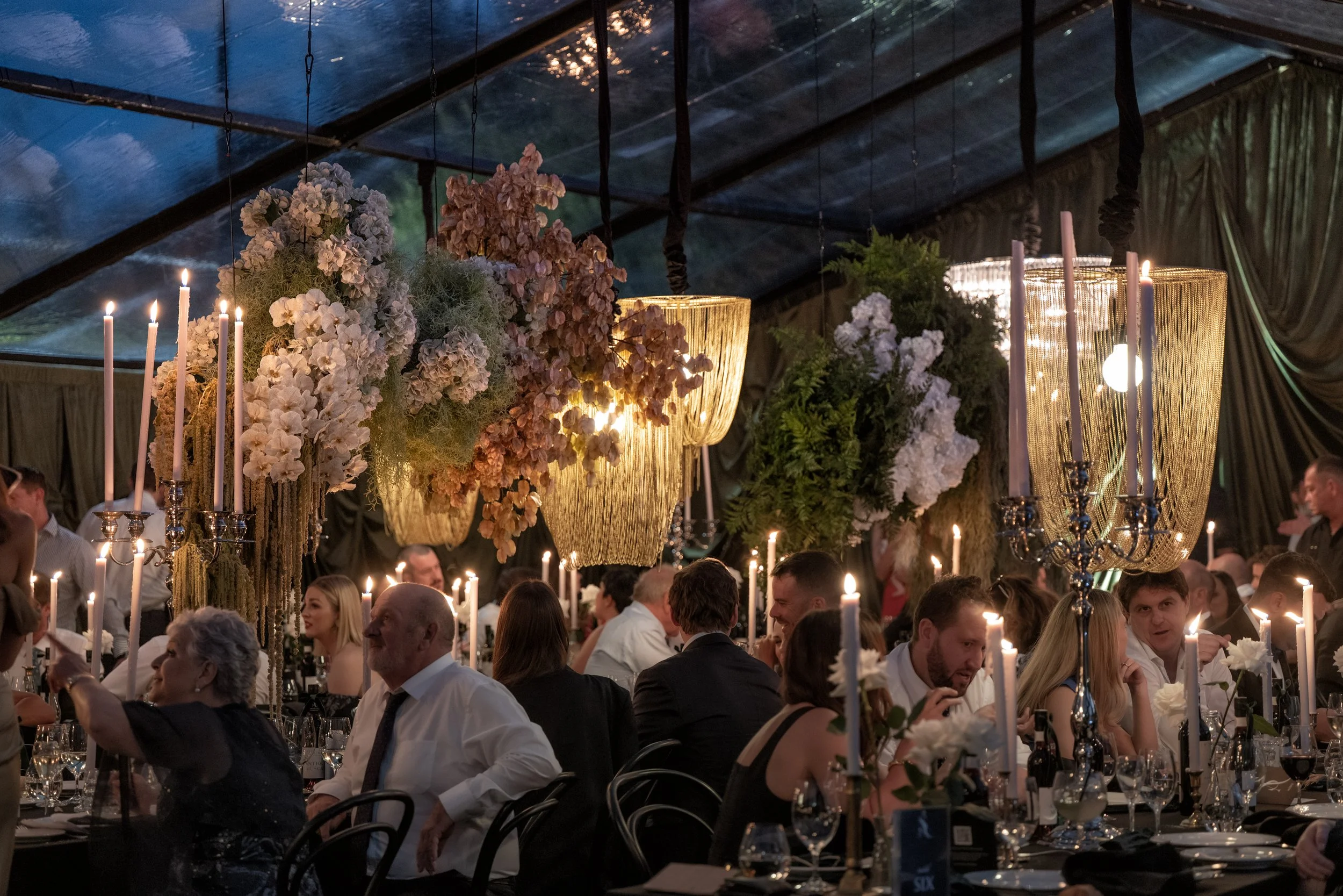 A large gathering of people seated at decorated tables inside a decorated tent with floral arrangements and hanging lights and candles.