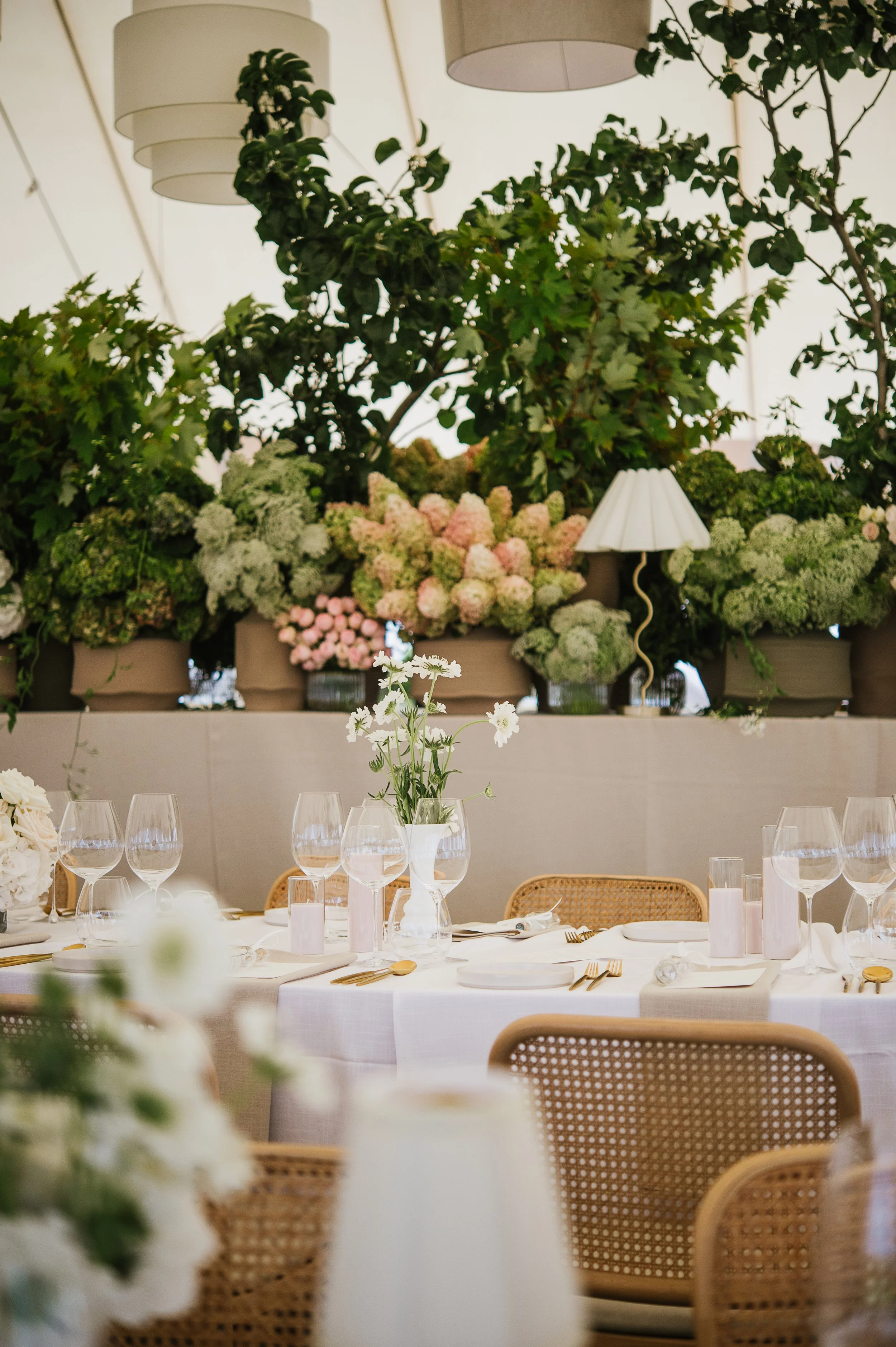 Elegant indoor dining setup with white tablecloths, floral centerpieces, wine glasses, gold flatware, and woven rattan chairs. In the background, there are large potted plants, overhead lamps, and floral arrangements on a ledge.