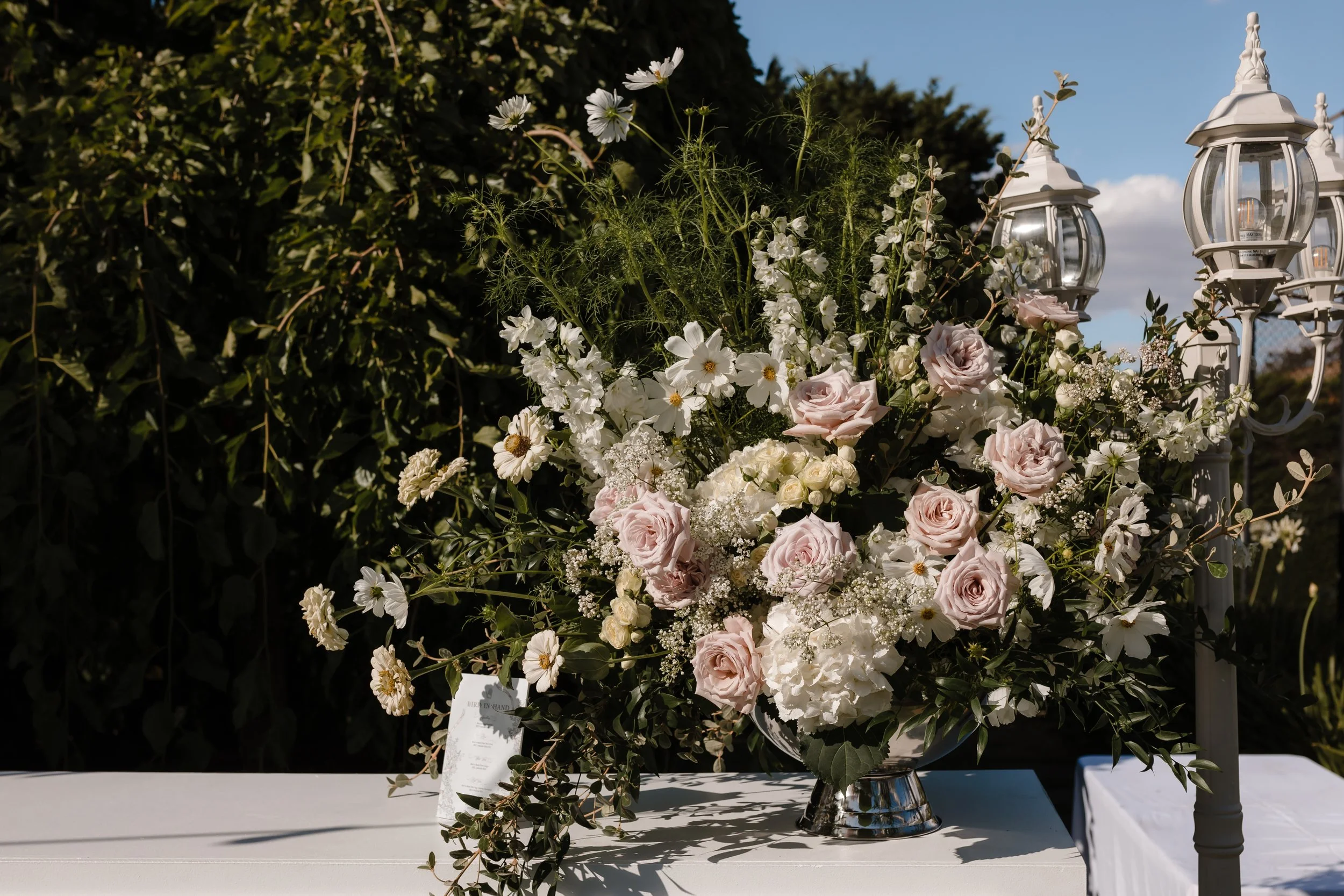 A large floral arrangement of pale pink roses, white daisies, white hydrangeas, and various greenery, placed on a white table with decorative lamp posts nearby, outdoors under a blue sky with some clouds.